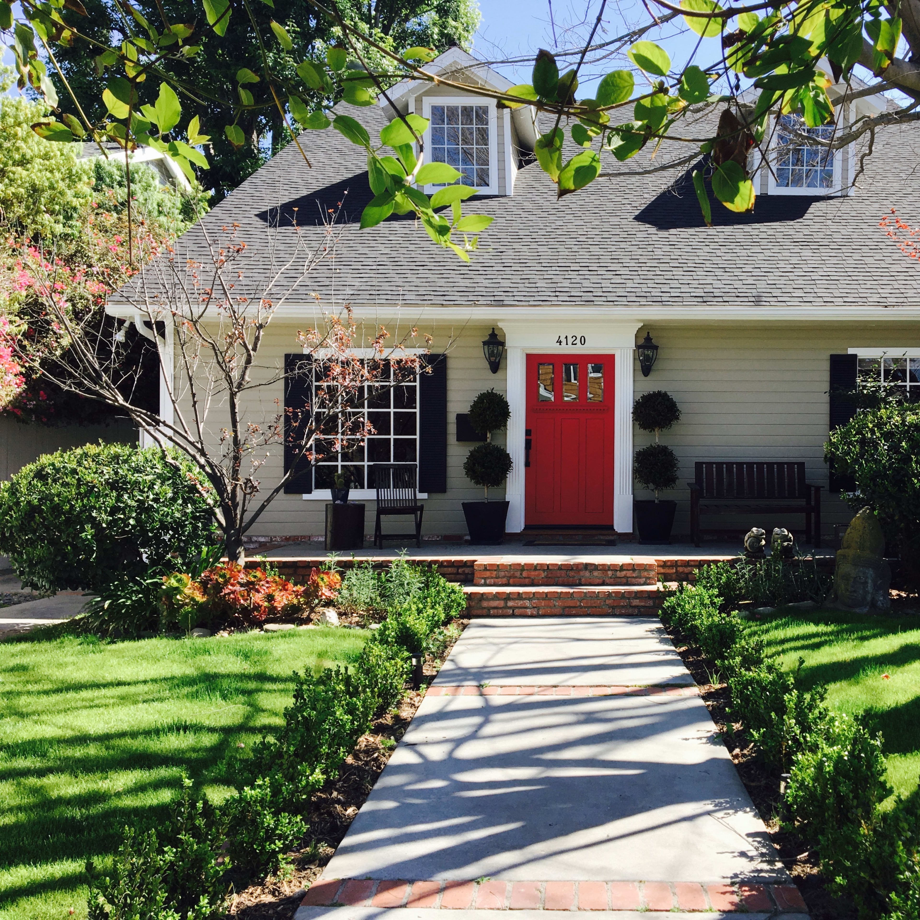 A single-story house with a gray exterior, a red front door, and neatly trimmed greenery lines a brick walkway leading to the entrance.