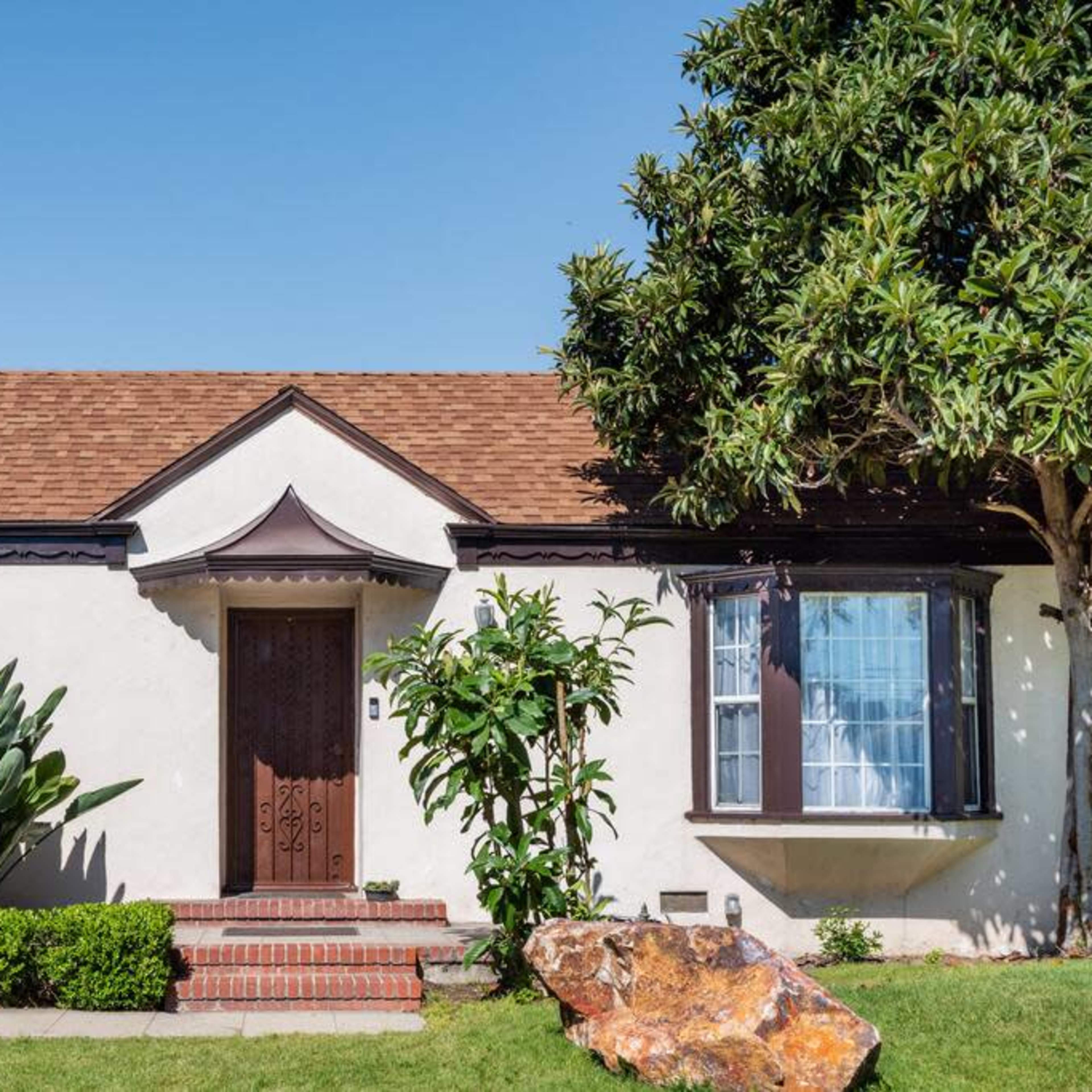 A single-story house with a brown shingle roof, a front porch, and landscaped greenery including shrubs and plants.