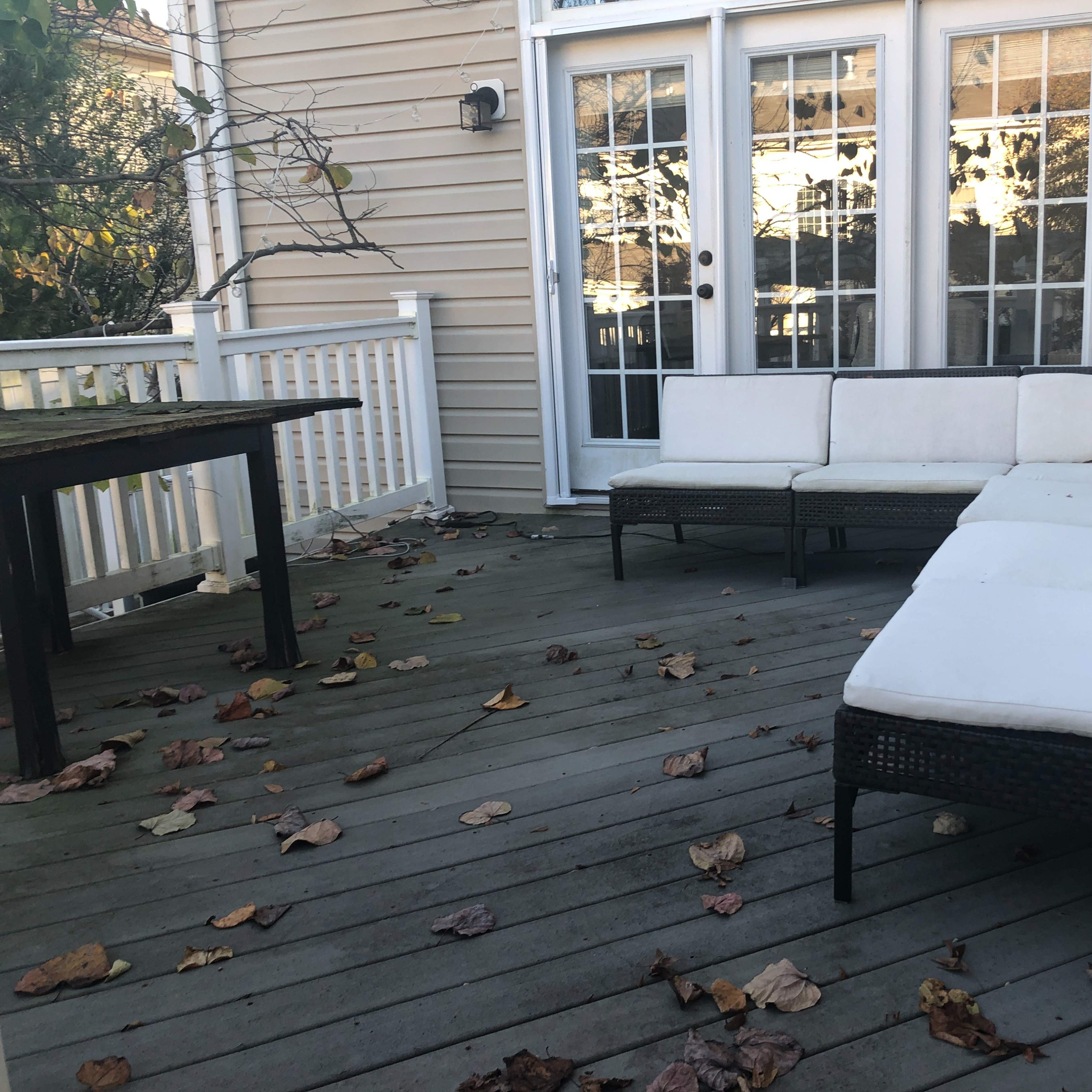 A white cushioned seating area is arranged on a wooden deck covered with fallen leaves, adjacent to a glass door leading inside.