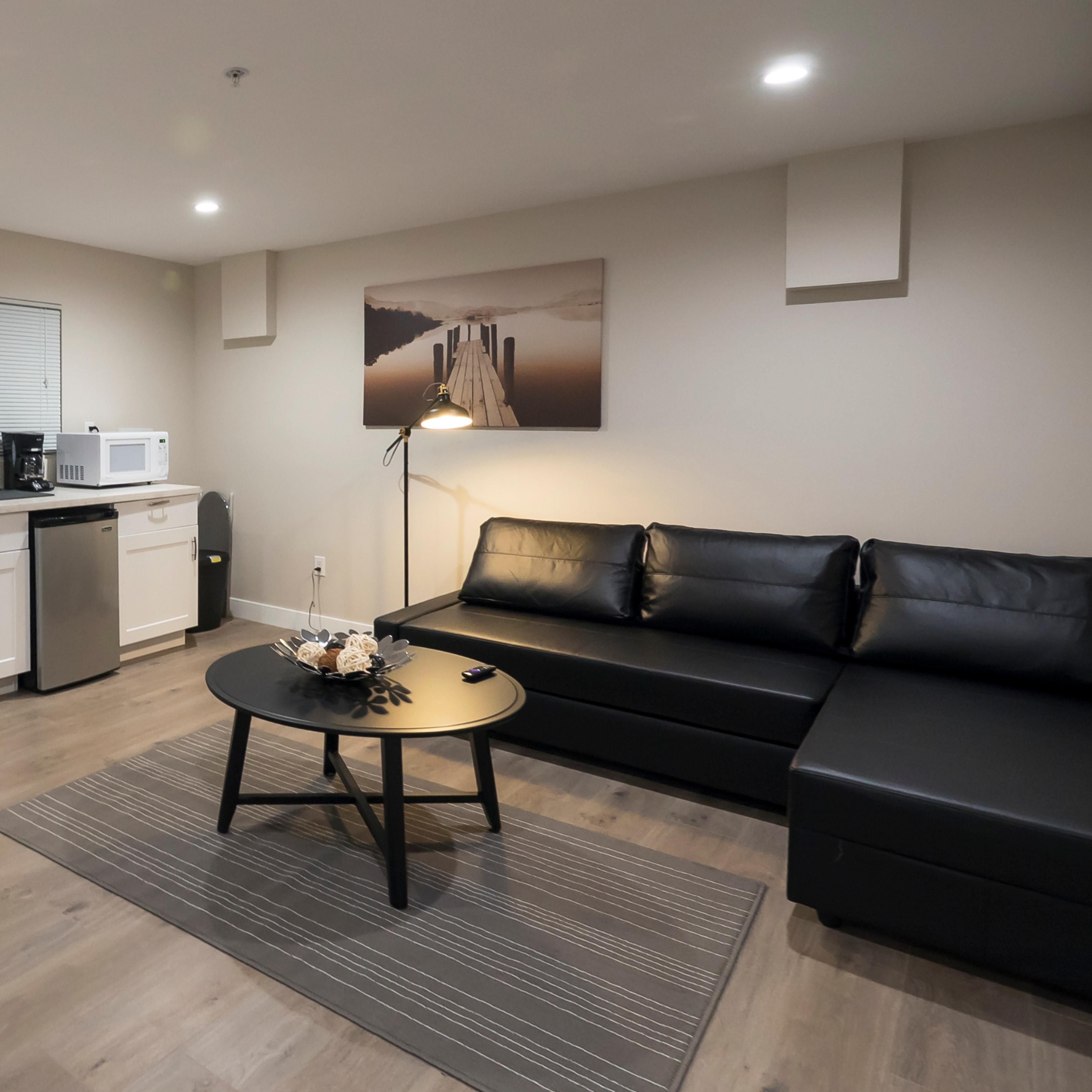 A modern living area with a black sectional sofa, a round coffee table, a television, and a kitchenette featuring a microwave and a refrigerator.