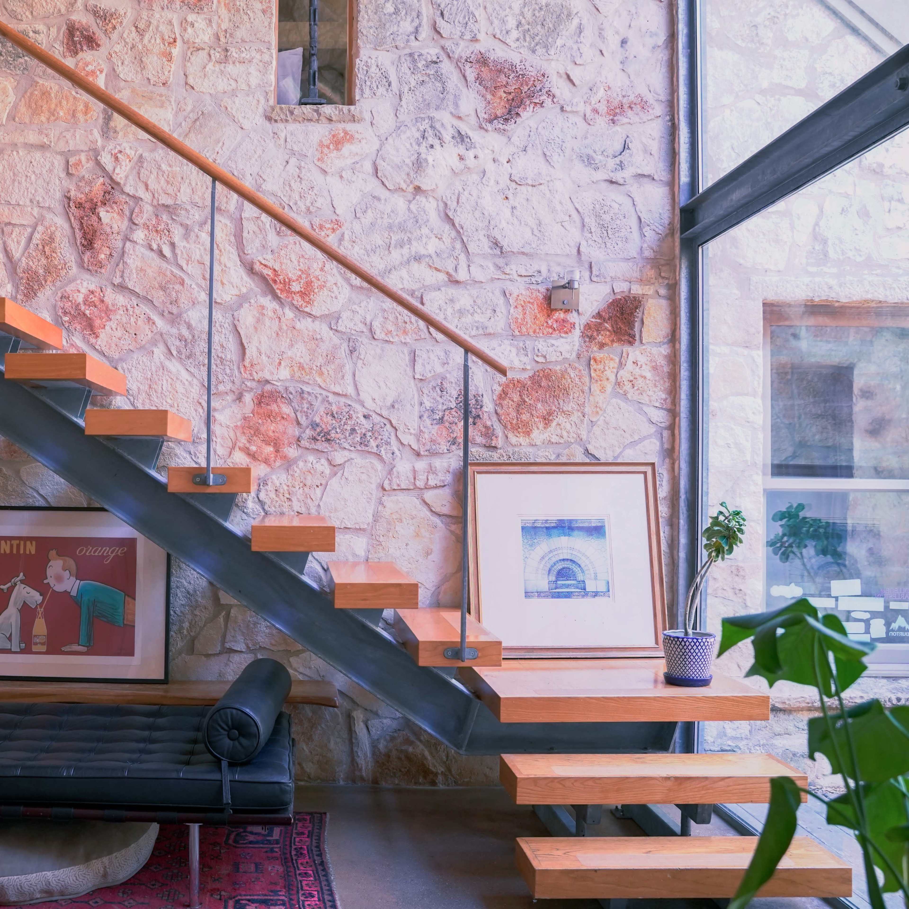 A wooden staircase with metal railings ascends beside a stone wall, featuring framed artwork and a plant on a side shelf.