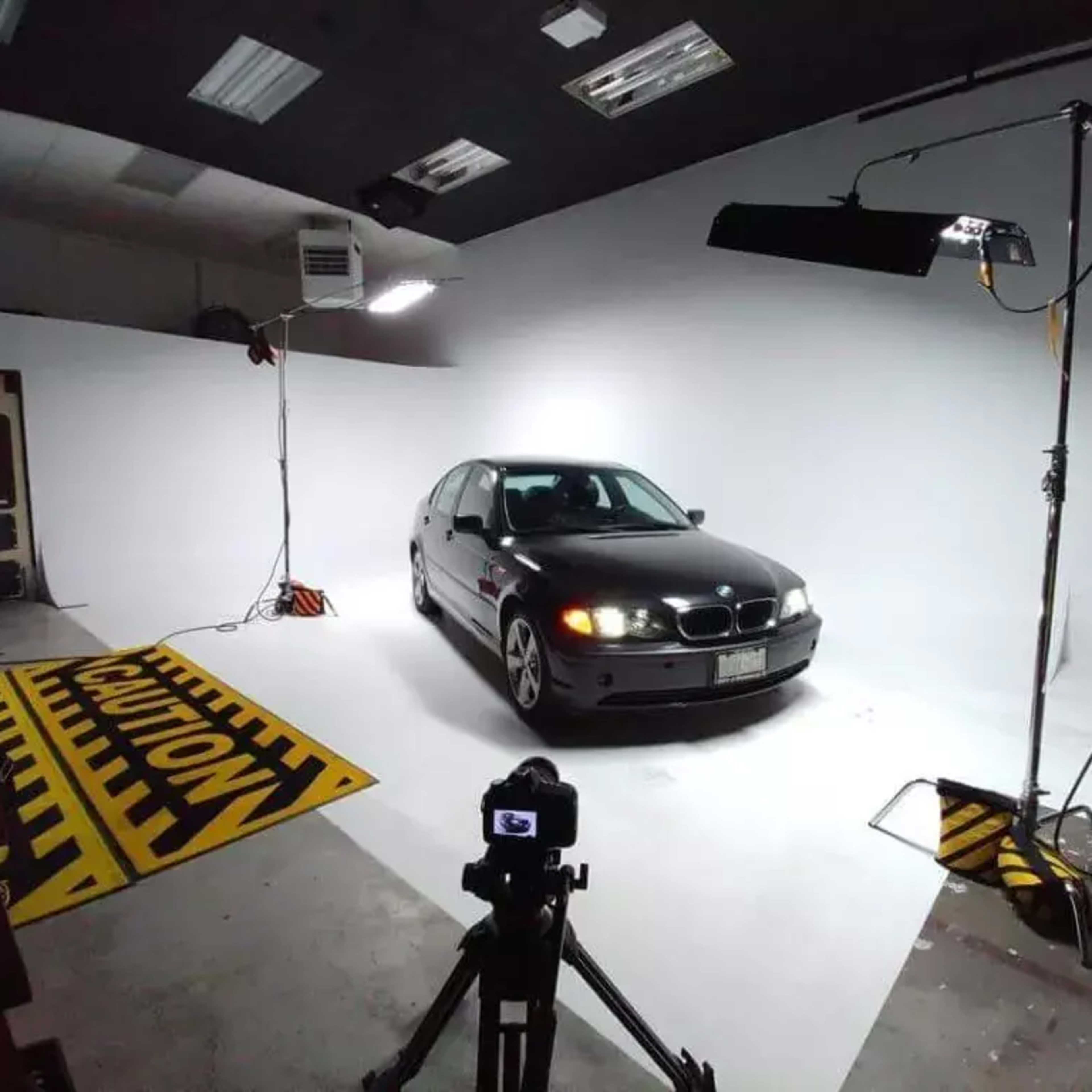 A black BMW sedan is positioned in a well-lit studio against a white backdrop, with lighting equipment and a camera set up for a photoshoot.