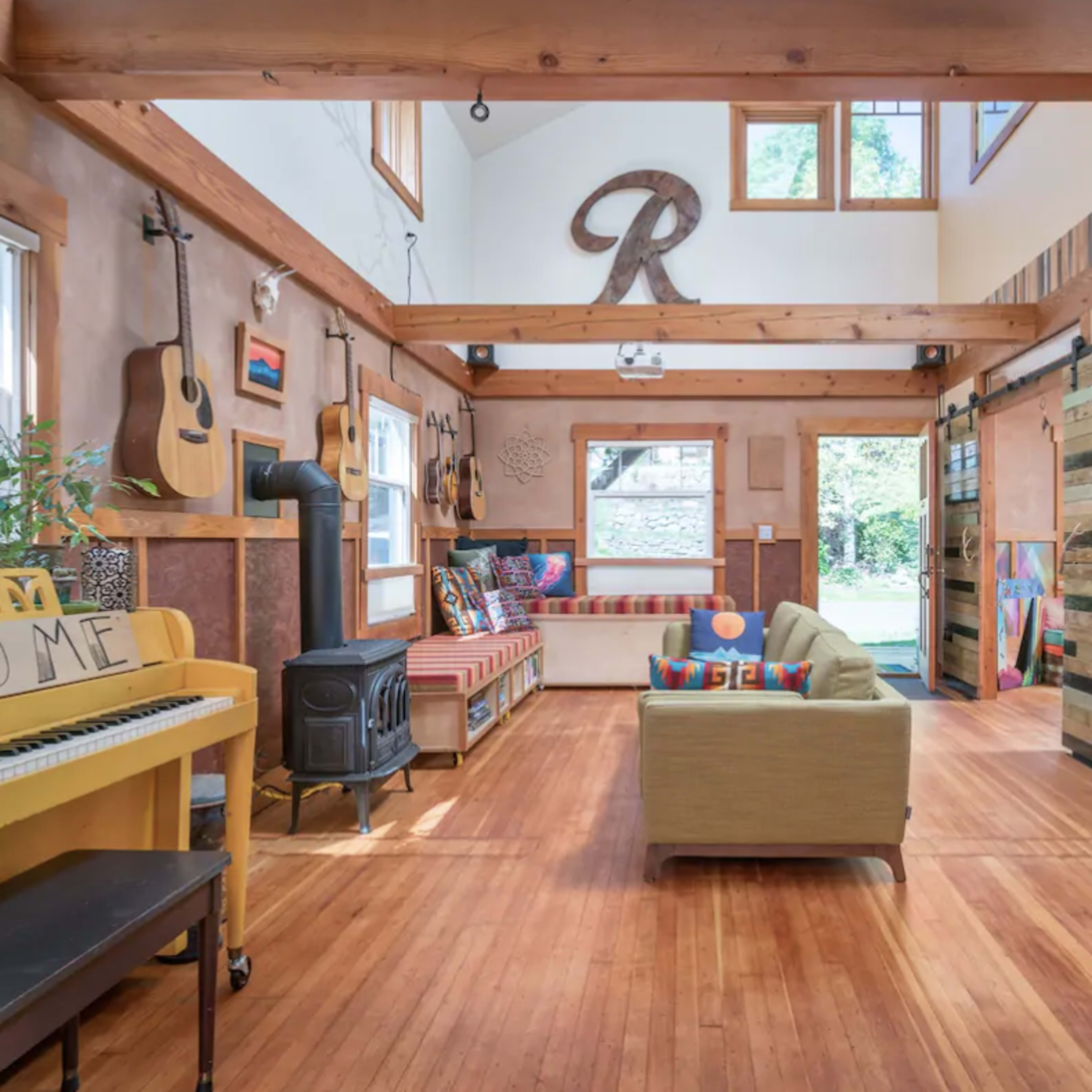 A brightly lit open living space featuring wooden beams, a piano, seating areas with colorful cushions, and various decorative guitars on the walls.