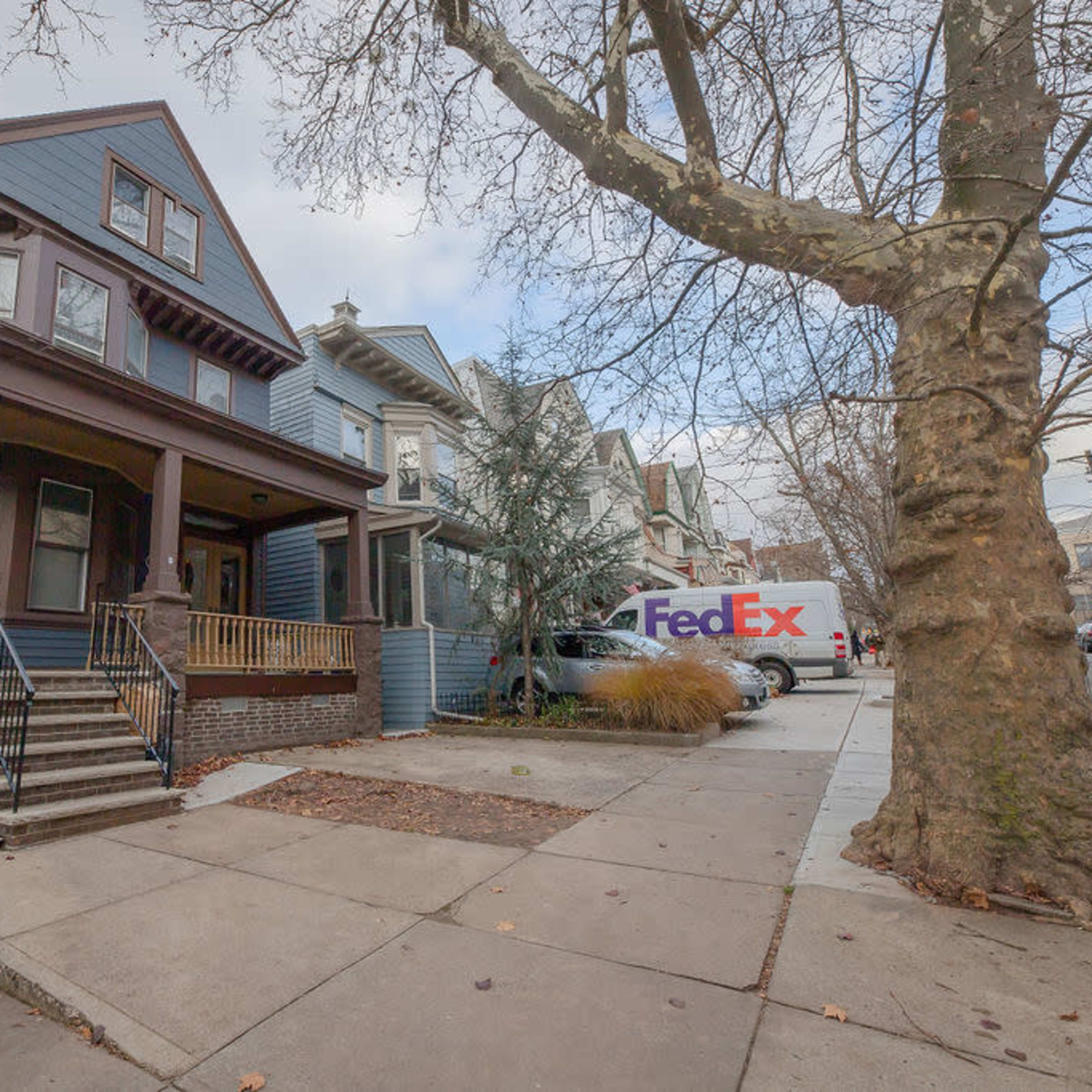 A blue two-story house with a porch stands beside a FedEx delivery truck parked on the street in an urban neighborhood.
