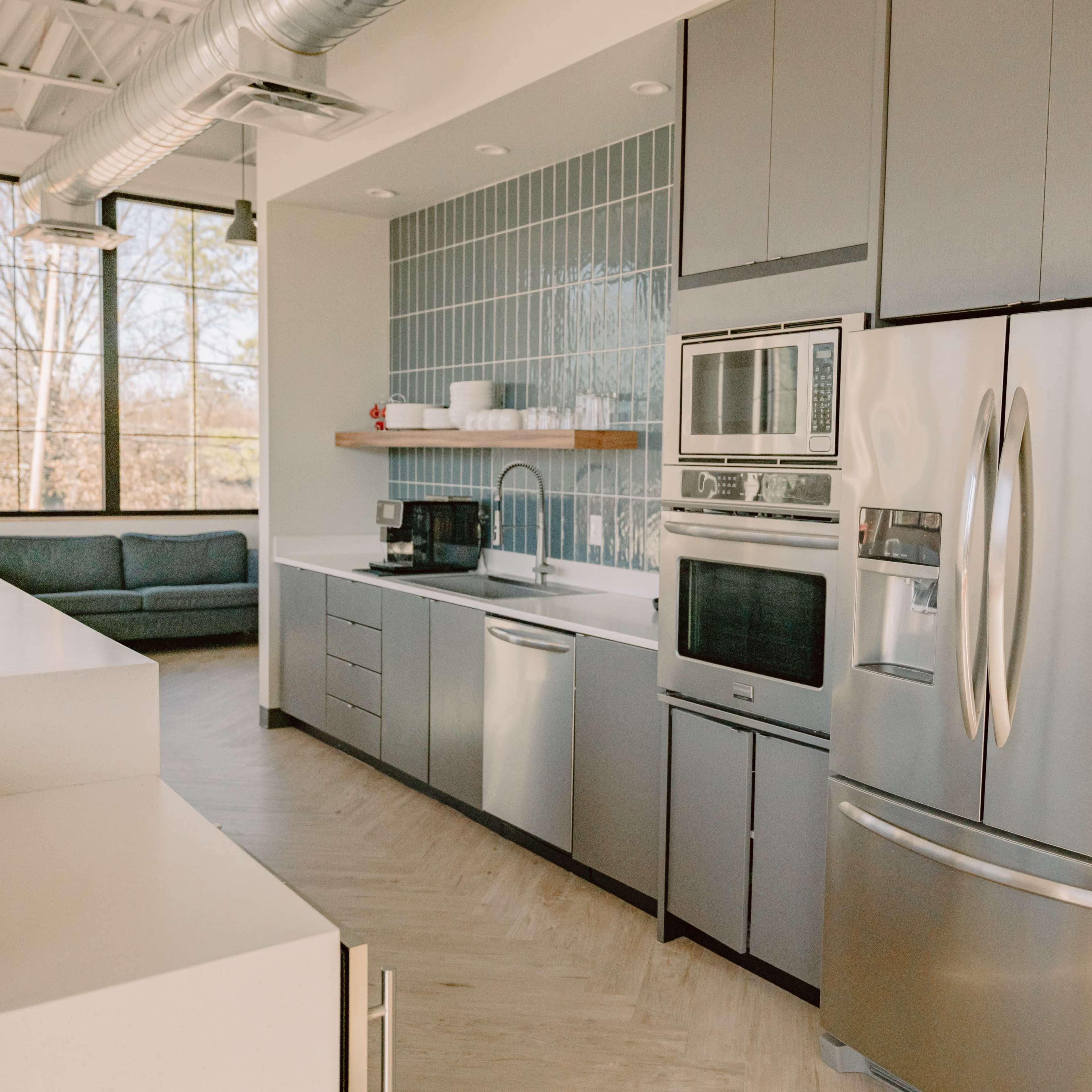 A modern kitchen featuring stainless steel appliances, grey cabinetry, and a window with a view of a couch and trees outside.