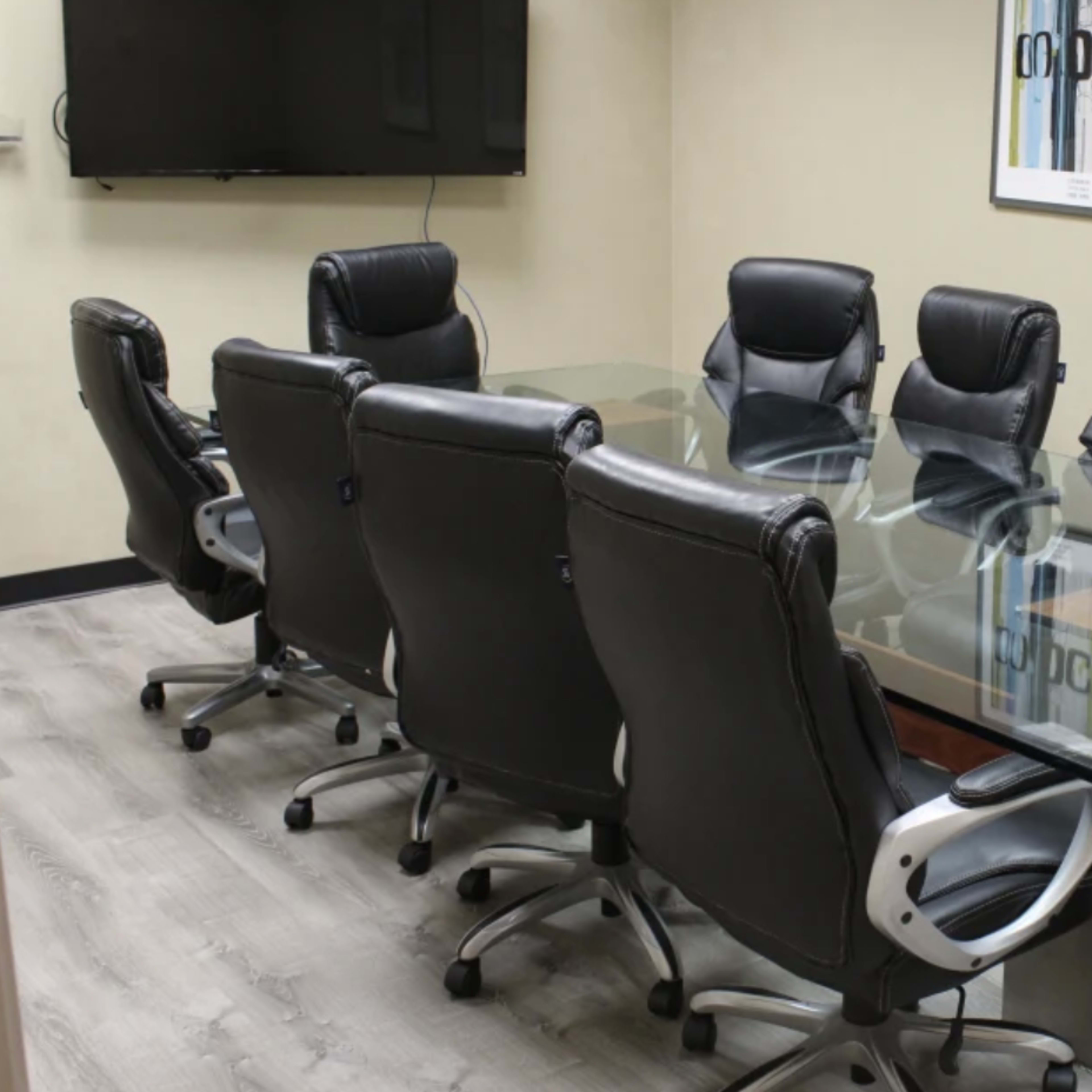 A conference room is set up with a glass table surrounded by eight black leather chairs, with a television mounted on the wall and framed artwork nearby.
