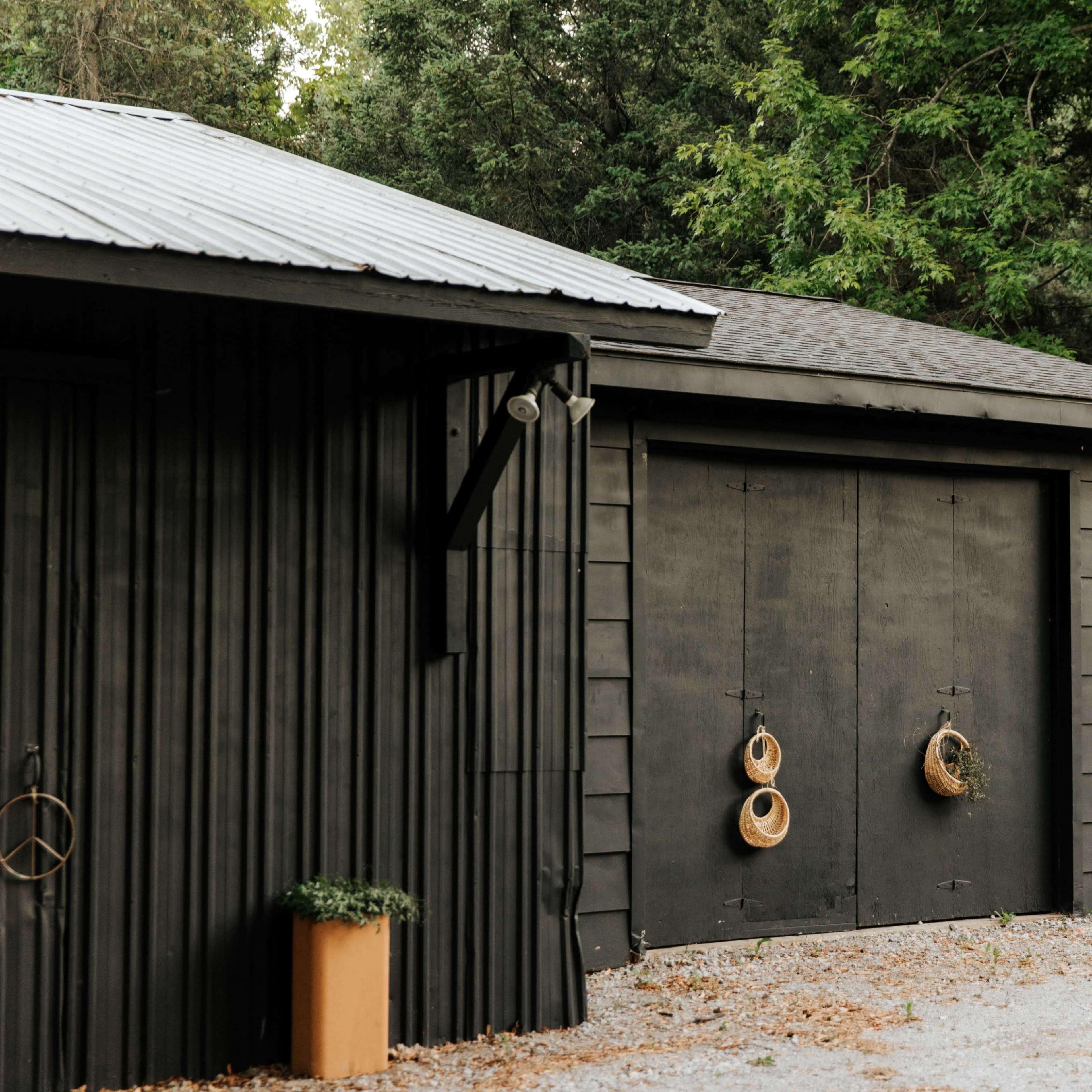 A black wooden garage features two large doors adorned with hanging baskets, situated alongside a gravel path and surrounded by greenery.