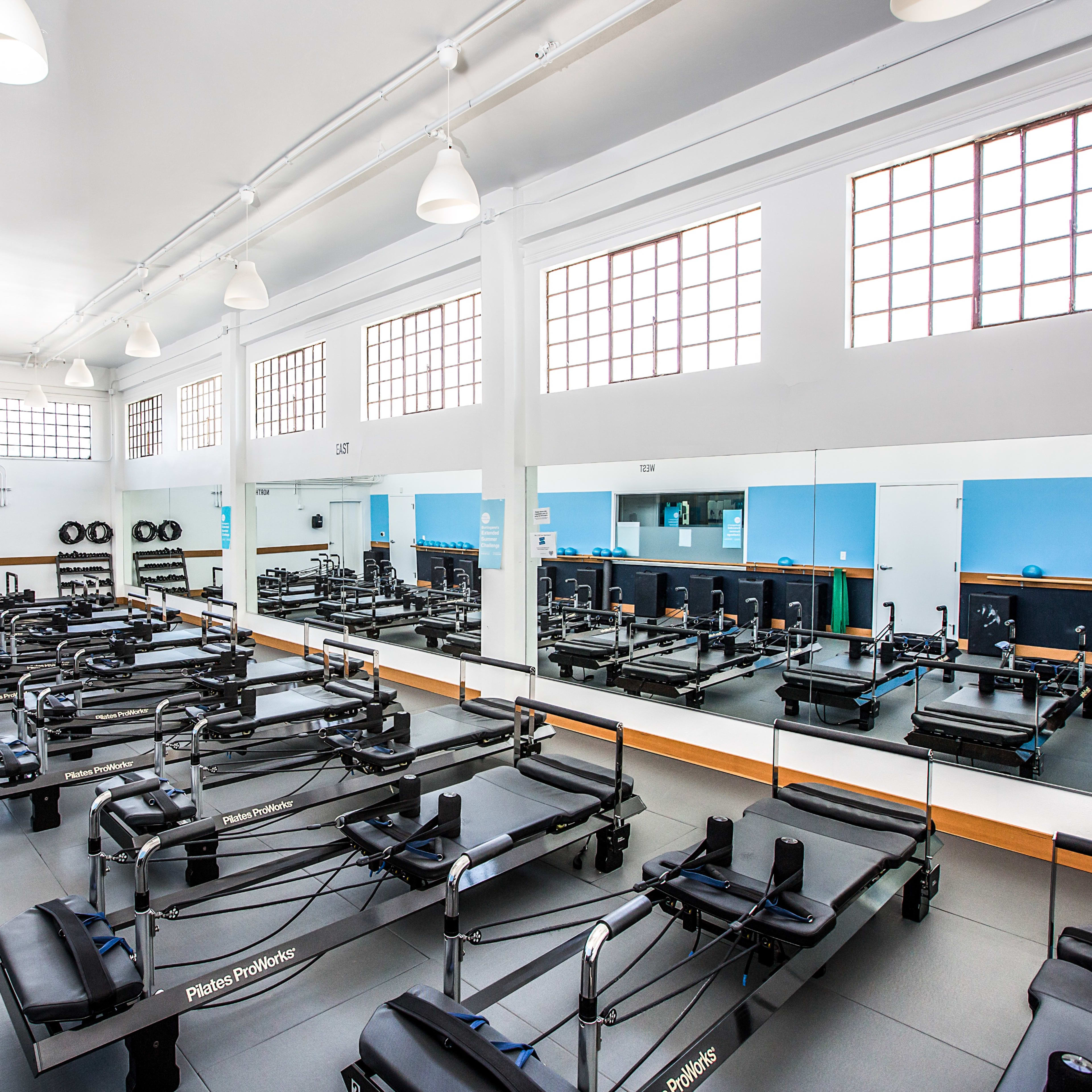 A bright, spacious Pilates studio filled with reformer machines arranged in neat rows, surrounded by large windows and mirrors.