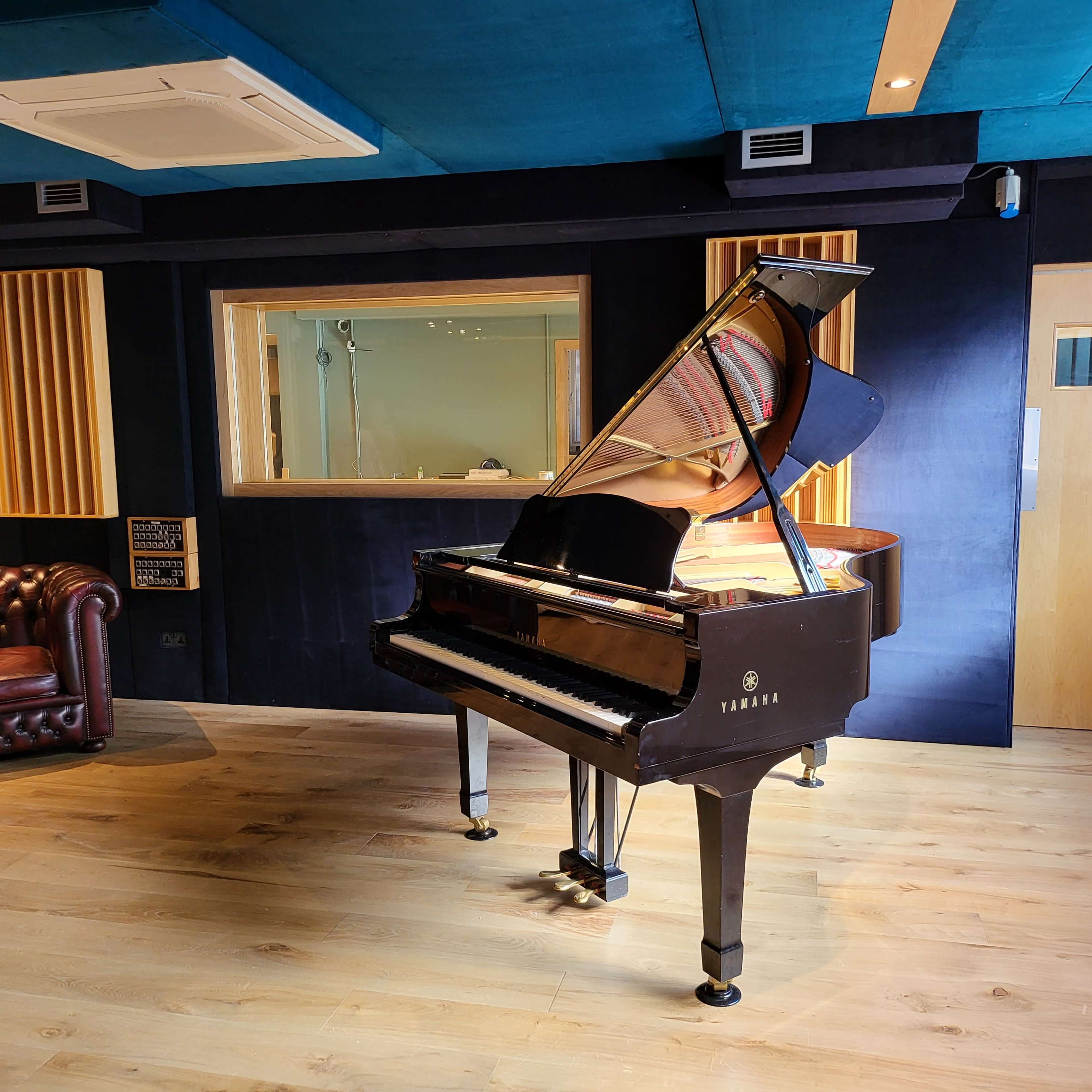 A grand piano is positioned in a recording studio with wooden flooring, a leather armchair nearby, and soundproofing panels on the walls.