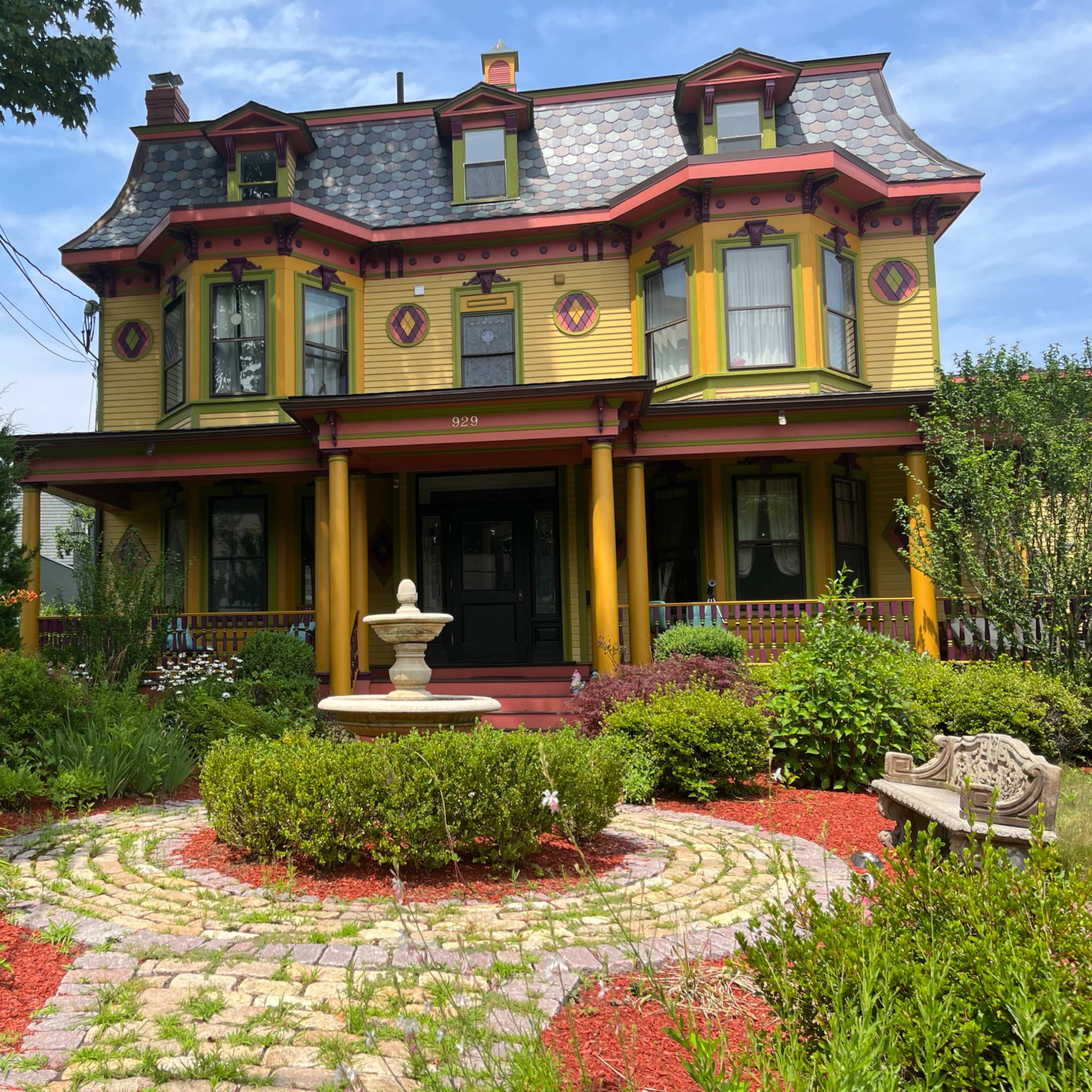 A Victorian-style house with yellow siding and red accents stands prominently in a landscaped garden featuring a stone fountain and circular stone pathway.