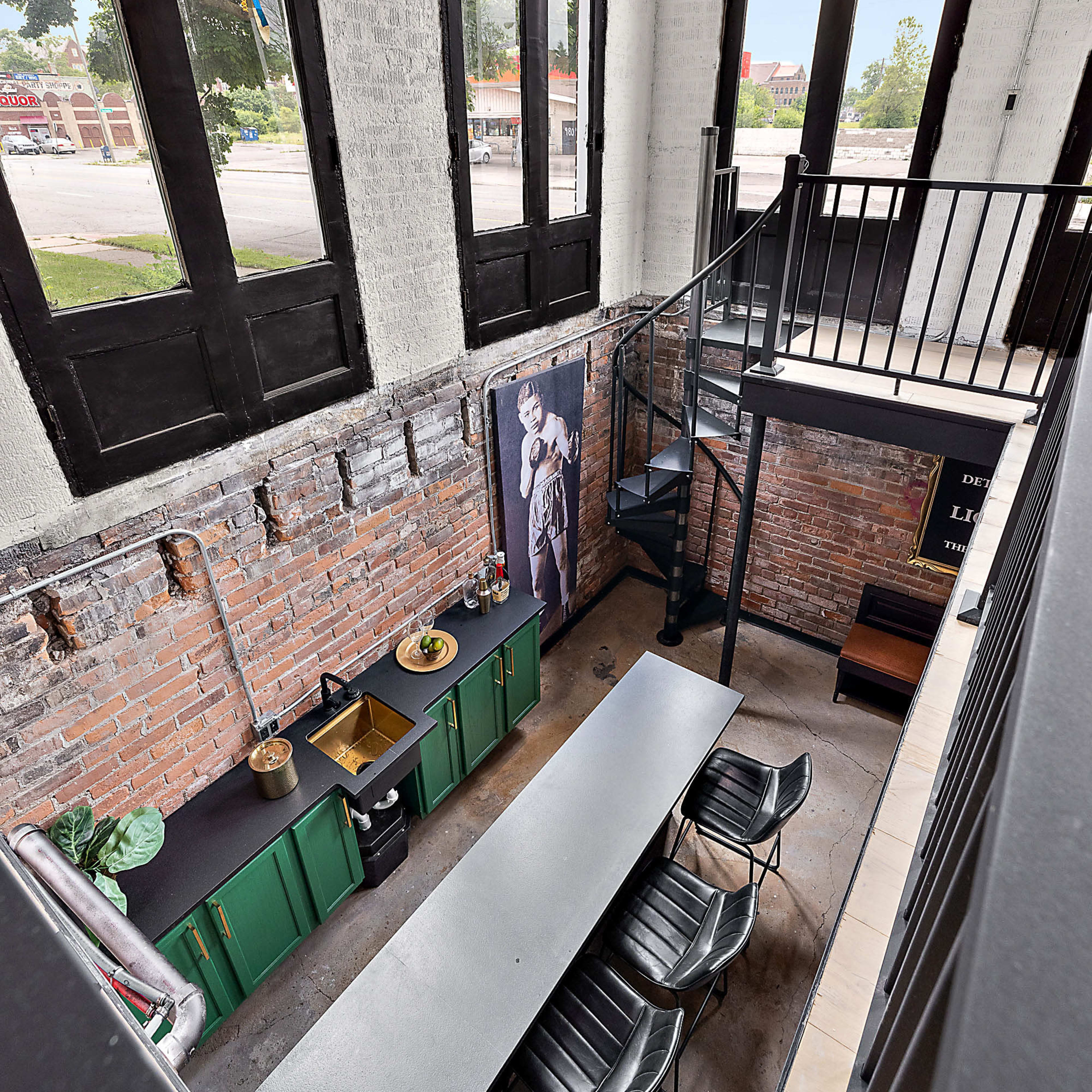An interior space with a high ceiling, featuring a long table set underneath large arched windows, and a green kitchen area alongside exposed brick walls.
