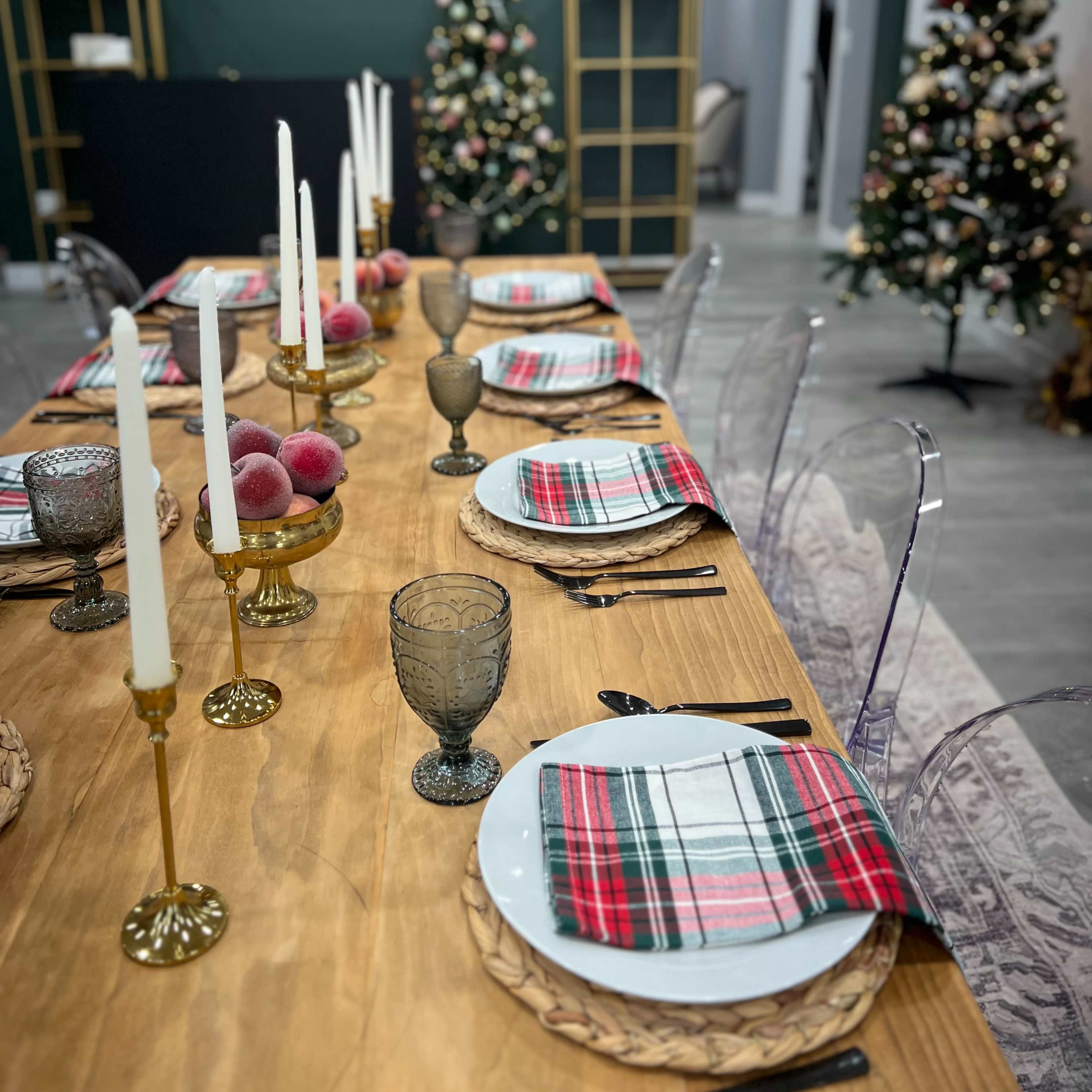 A holiday dining table is set with plaid plates and glassware, flanked by two decorated Christmas trees in a room with dark green walls.