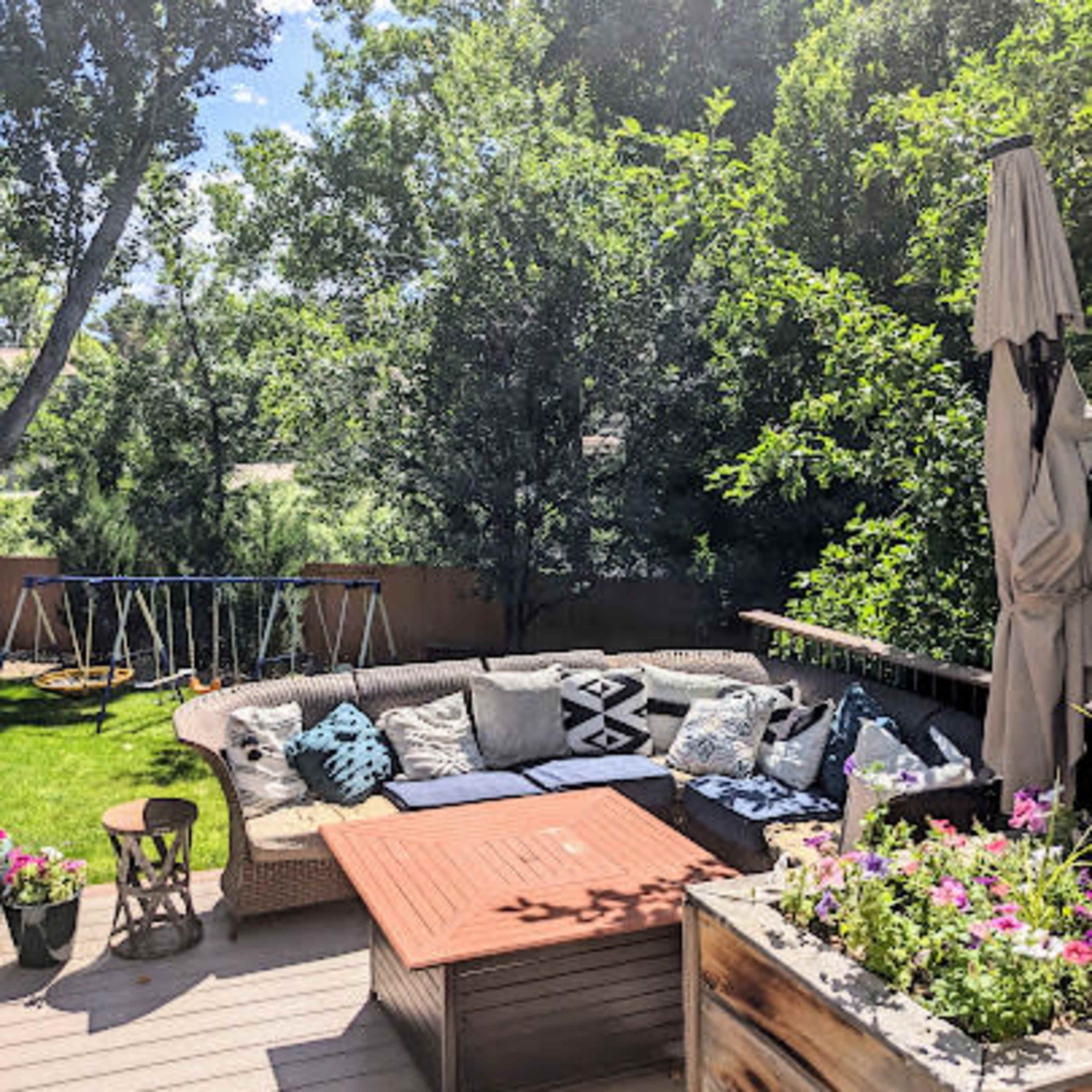 A patio seating area is arranged with a wicker couch and a wooden table, surrounded by greenery and potted plants.