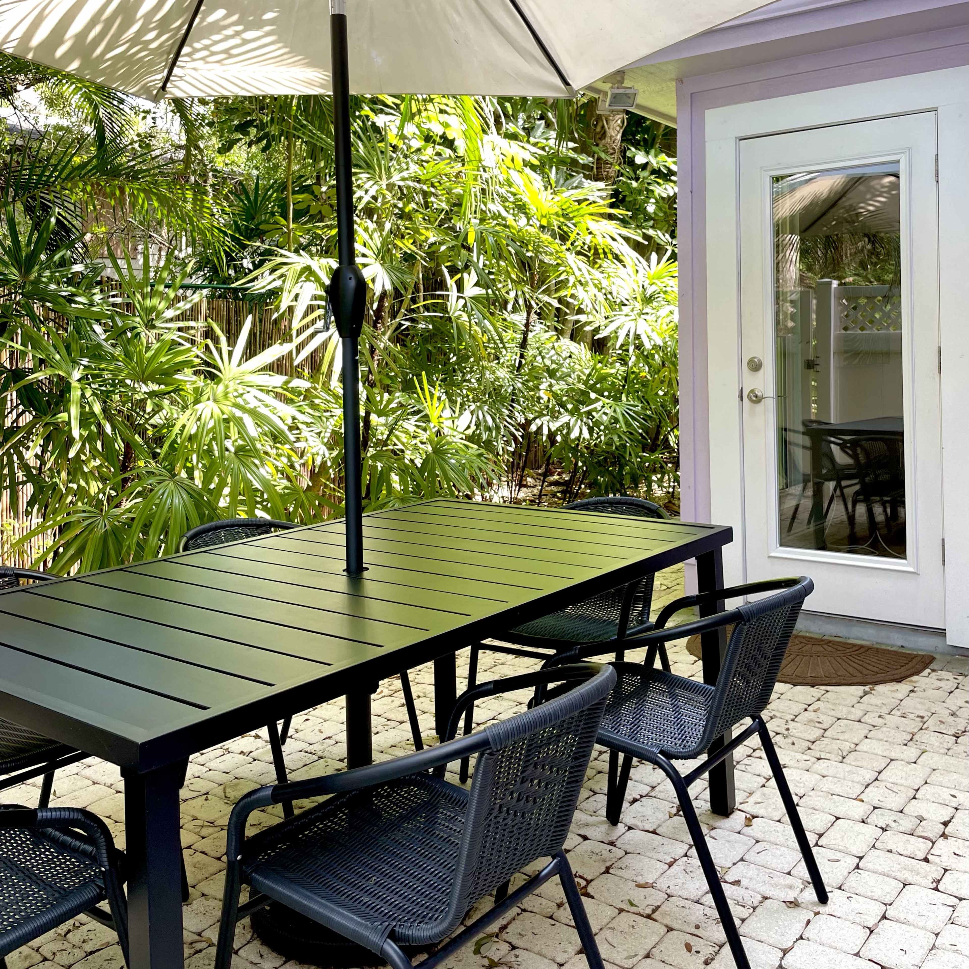 A metal dining table with six black chairs sits beneath a large umbrella in a patio surrounded by lush greenery and a small building with a mirrored door.