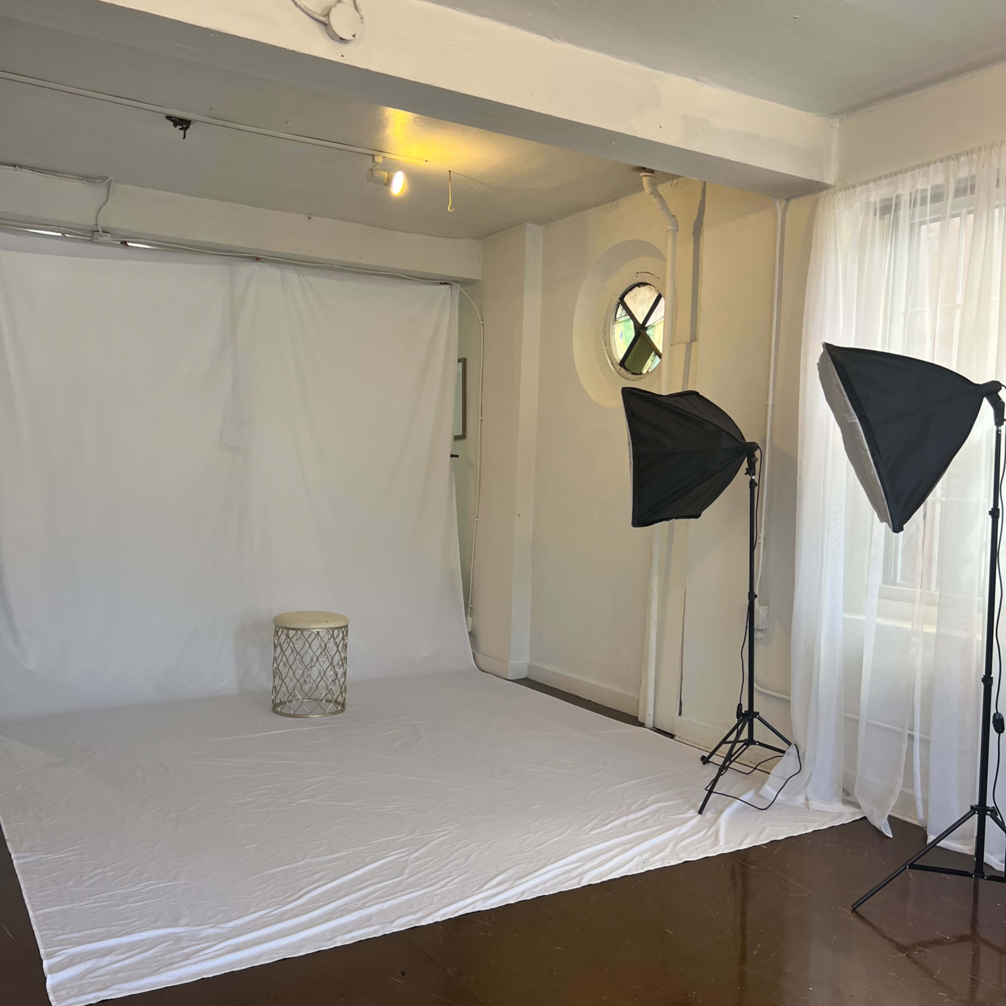A photography studio with a white backdrop, a small round stool, and two lighting setup stands.