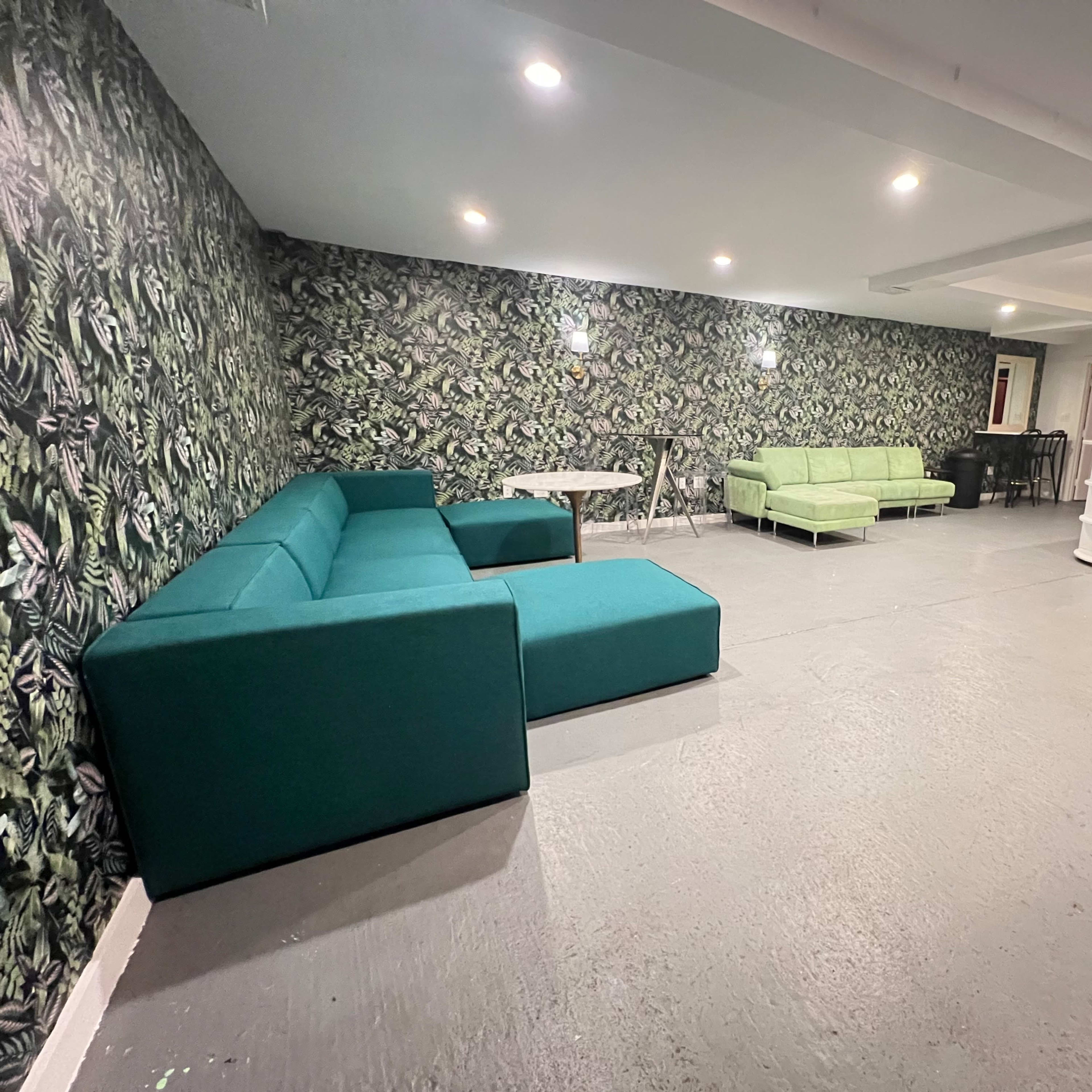 A room with a patterned green wallpaper, featuring three teal sofas arranged around a table, along with a light-colored seating area and a closet.