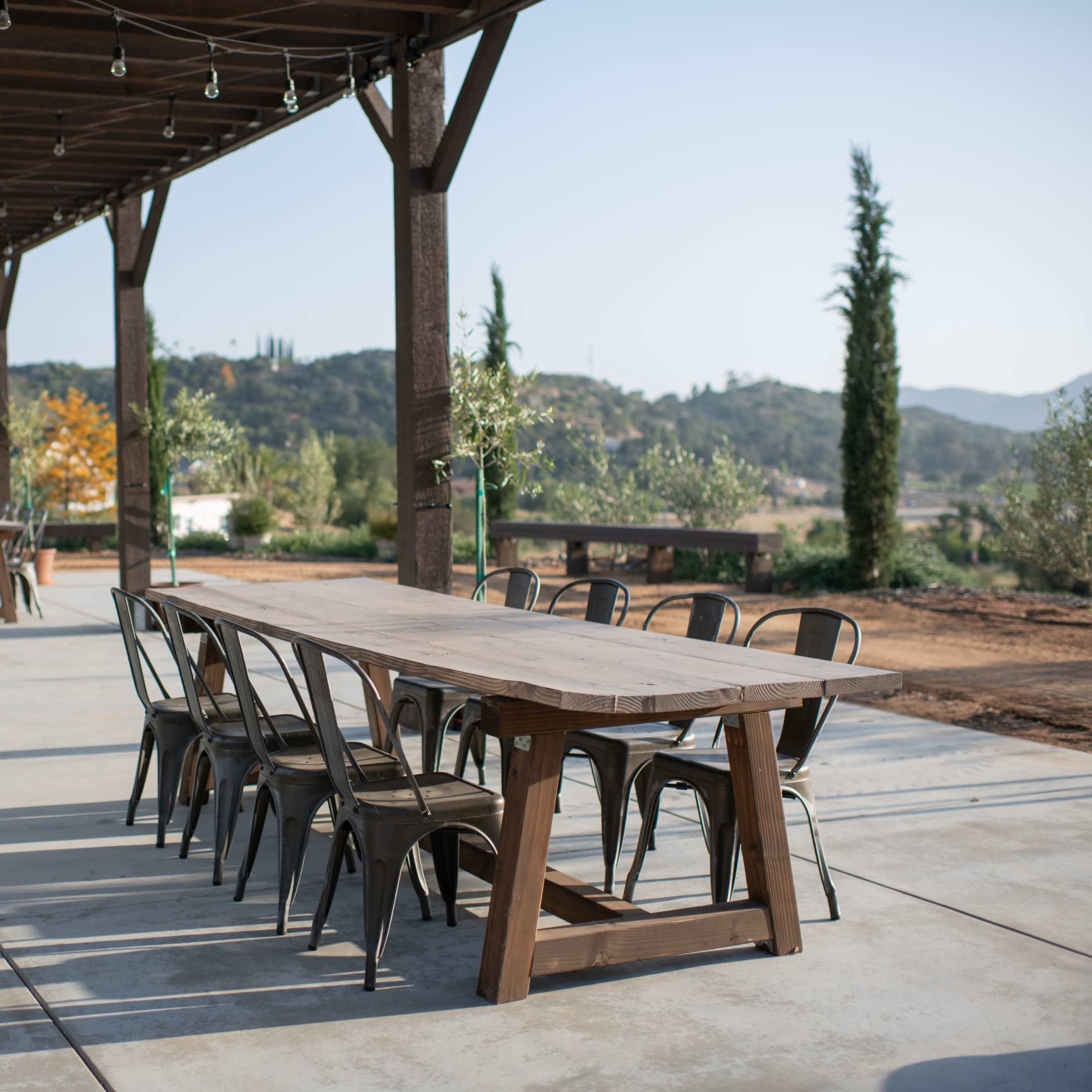 A spacious outdoor dining area features long wooden tables surrounded by metal chairs under a covered structure.
