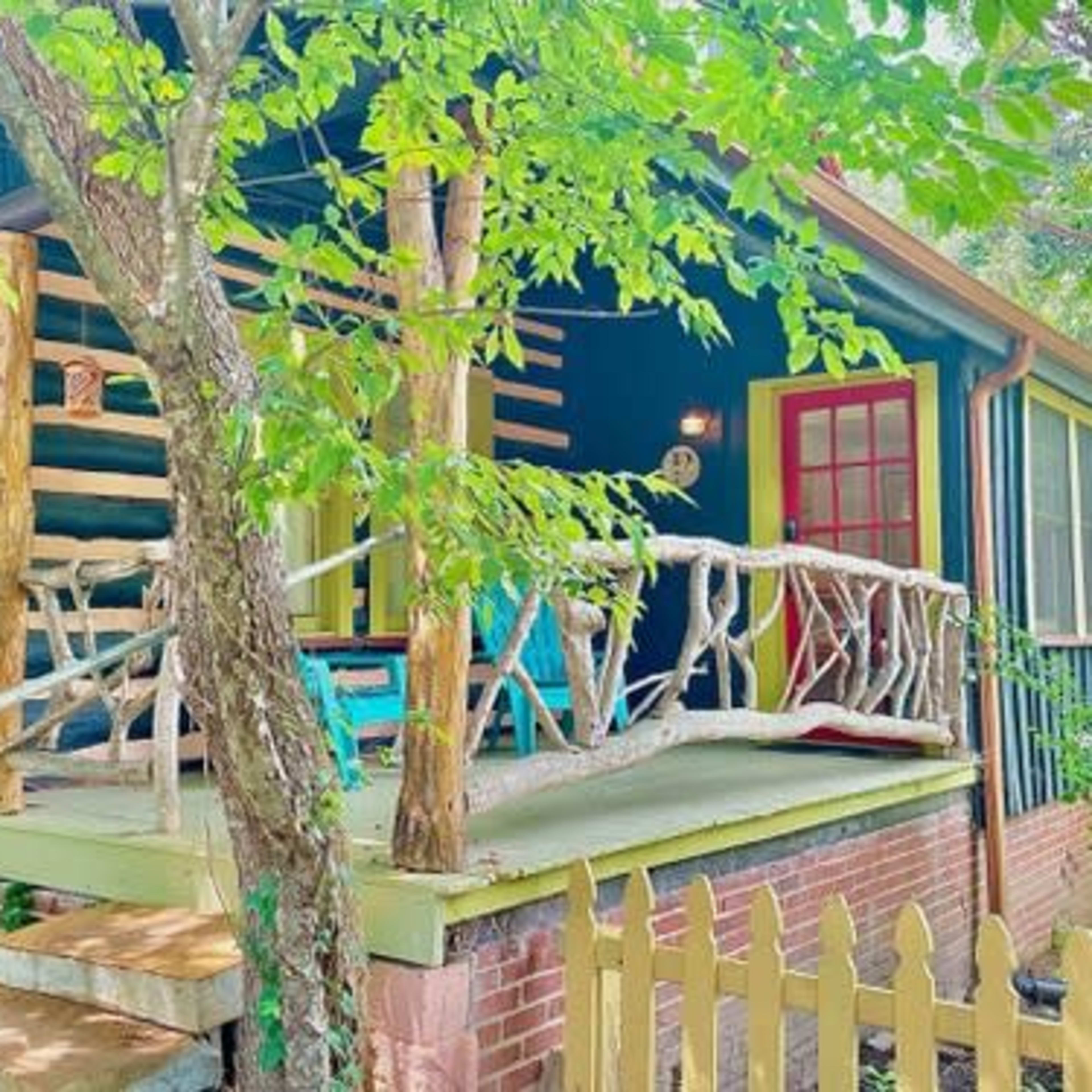A small log cabin with a porch features a red door, colorful furniture, and a wooden railing surrounded by greenery.