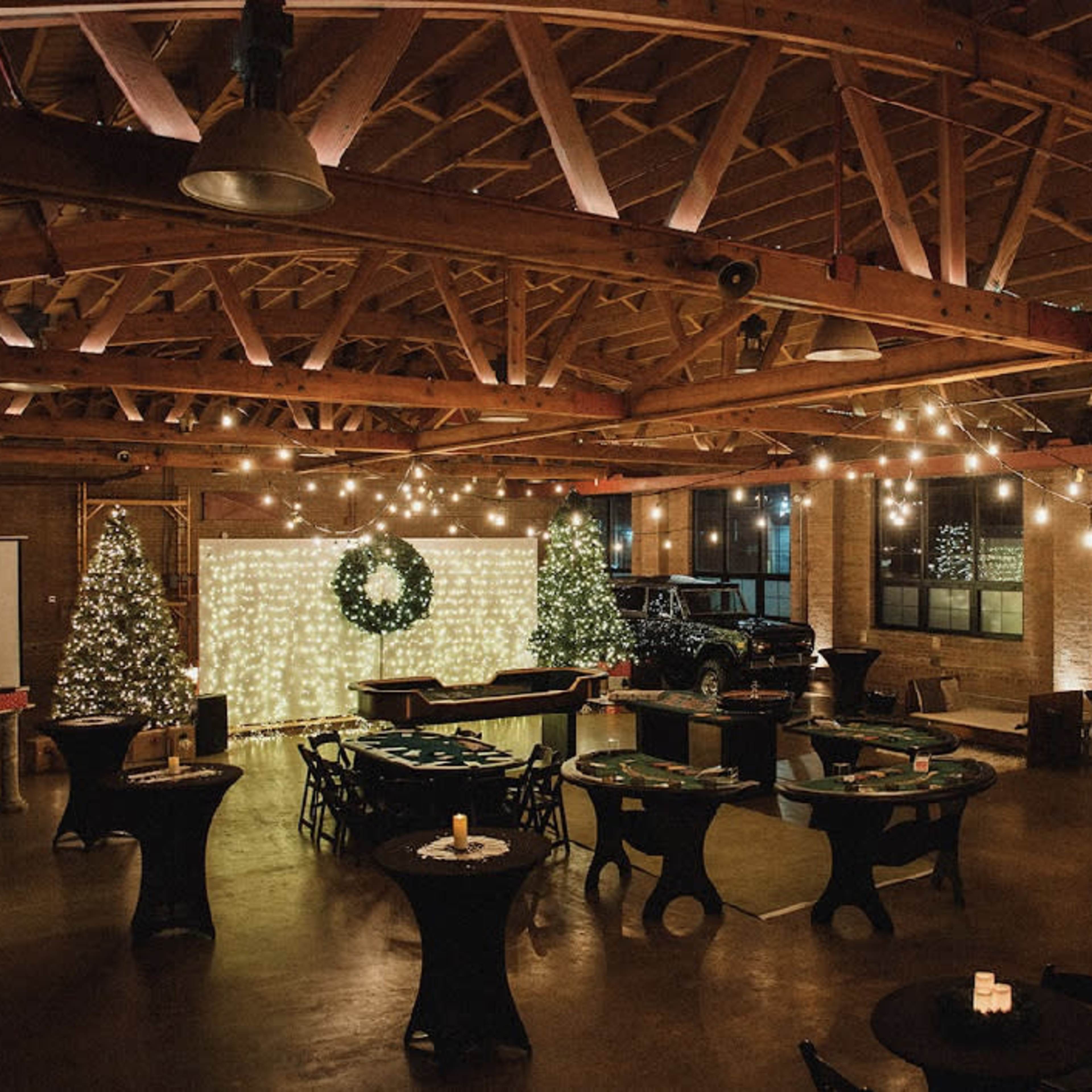 A spacious event venue with wooden beams, decorated with string lights and a festive backdrop, featuring tables arranged for a gathering.