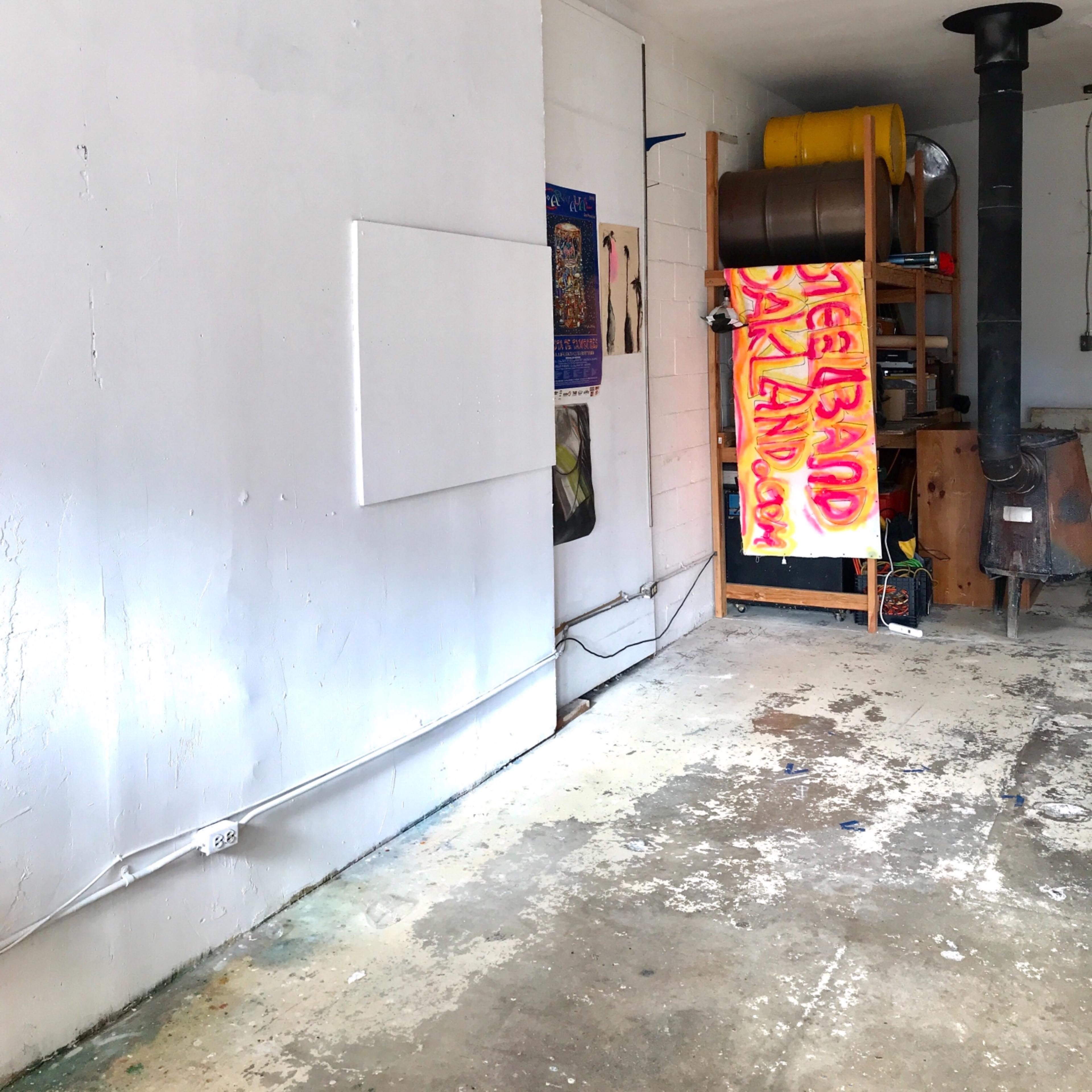 An empty garage interior with white walls, a wooden shelf displaying barrels, and a colorful sign hanging nearby.