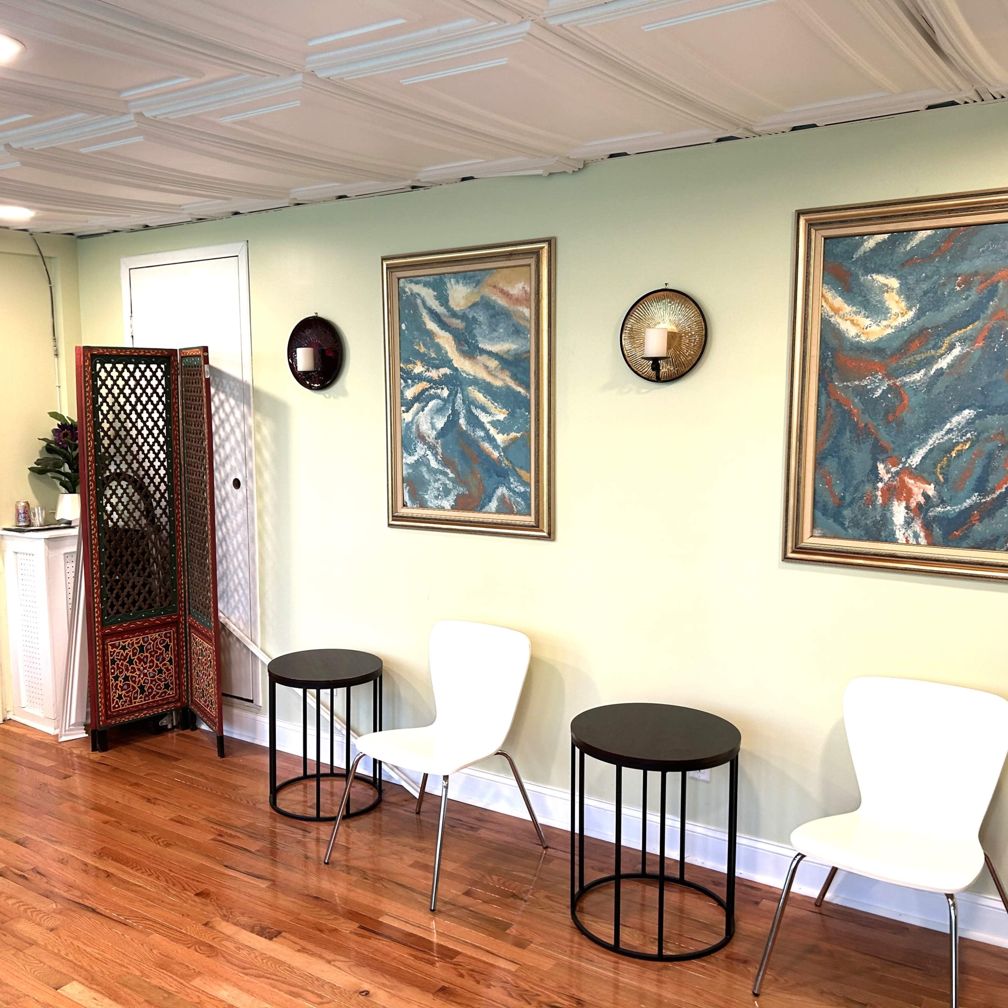 A room with light green walls, two framed abstract paintings, a lattice room divider, three chairs (two white and one black), and a door leading outside.