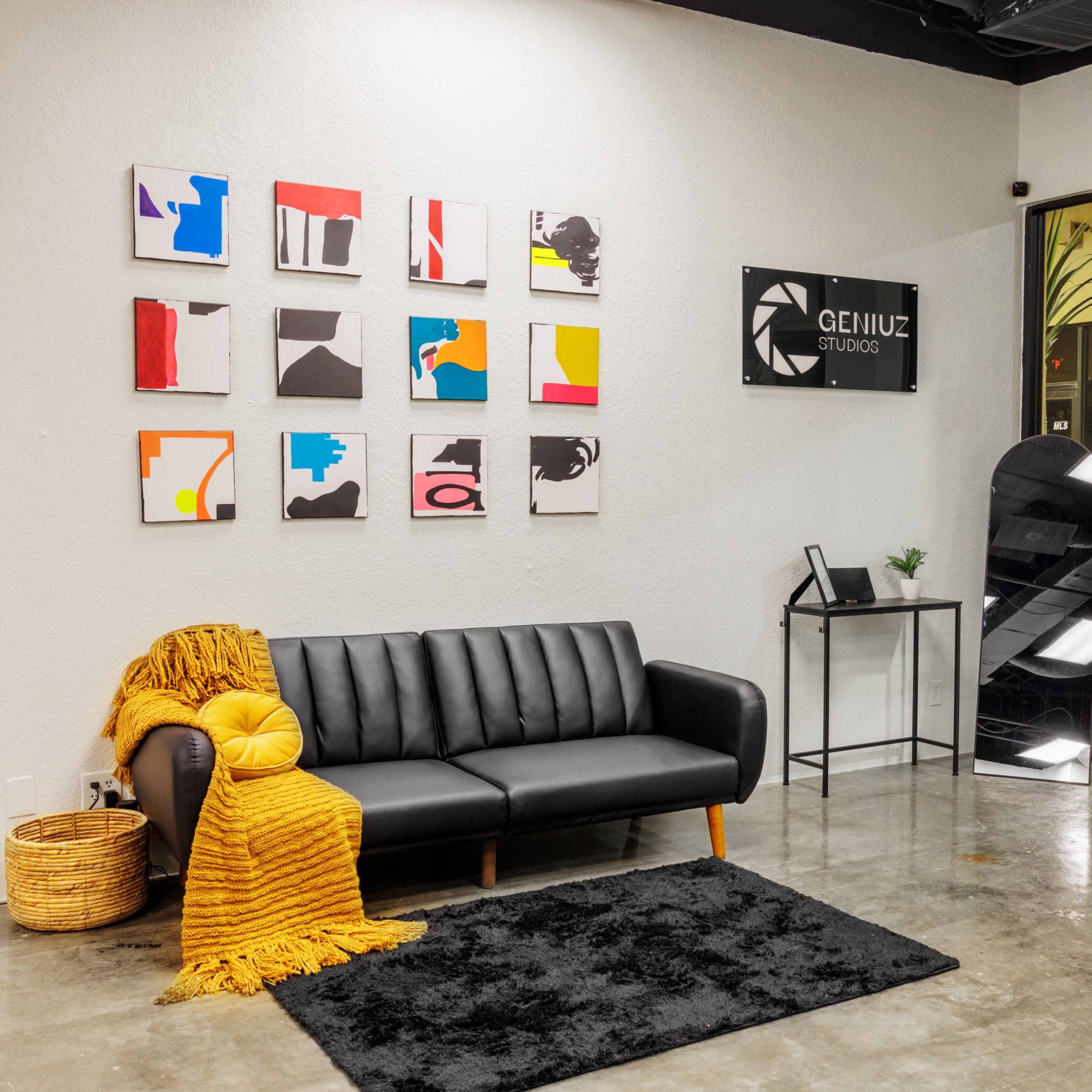 A modern lobby area with a black sofa, a yellow throw blanket, a textured black rug, and abstract art on the wall, along with a sign for "GENIUZ STUDIO."