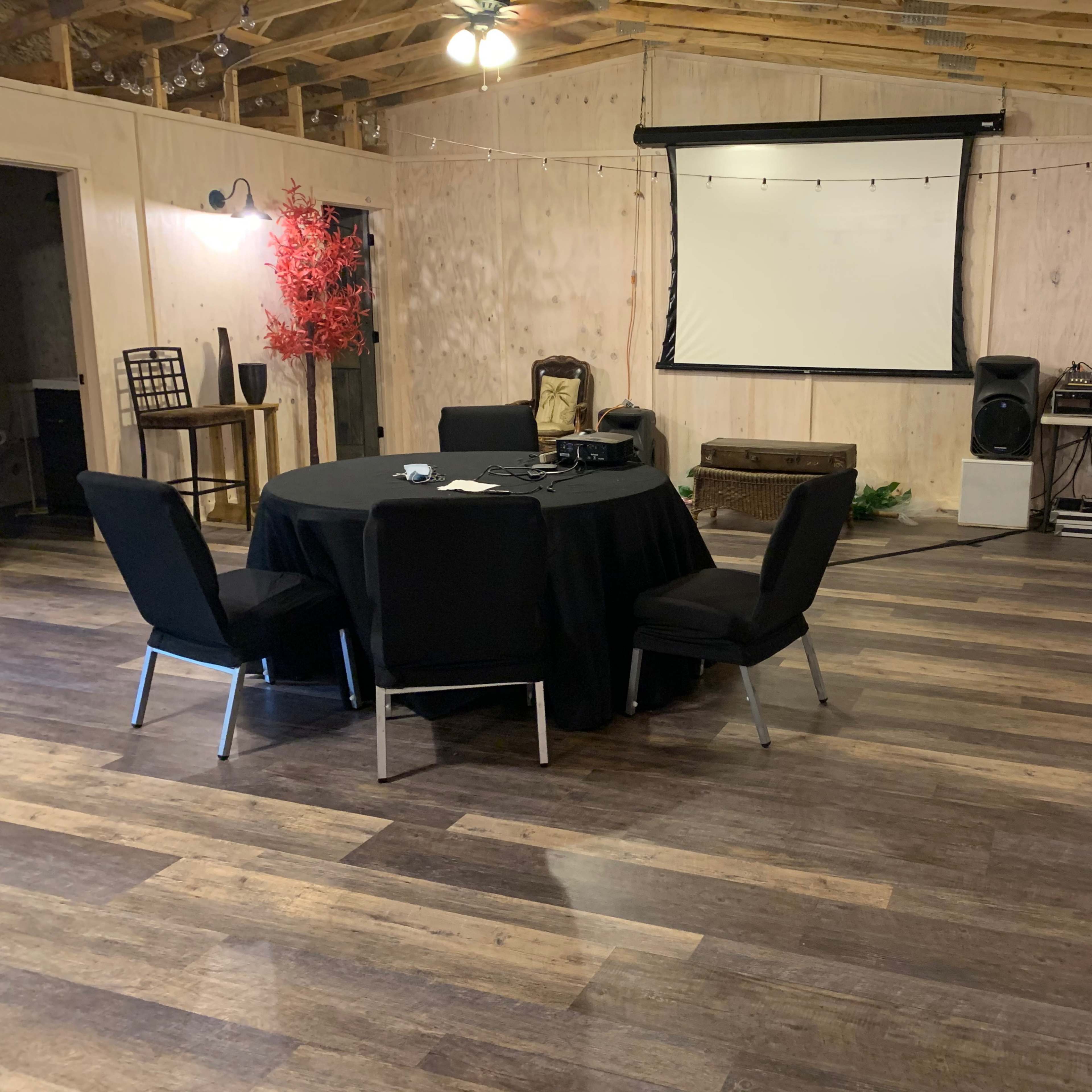 The interior of a room features a round table with dark chairs, a projector screen, and wooden flooring, accompanied by decorative elements and audio equipment.