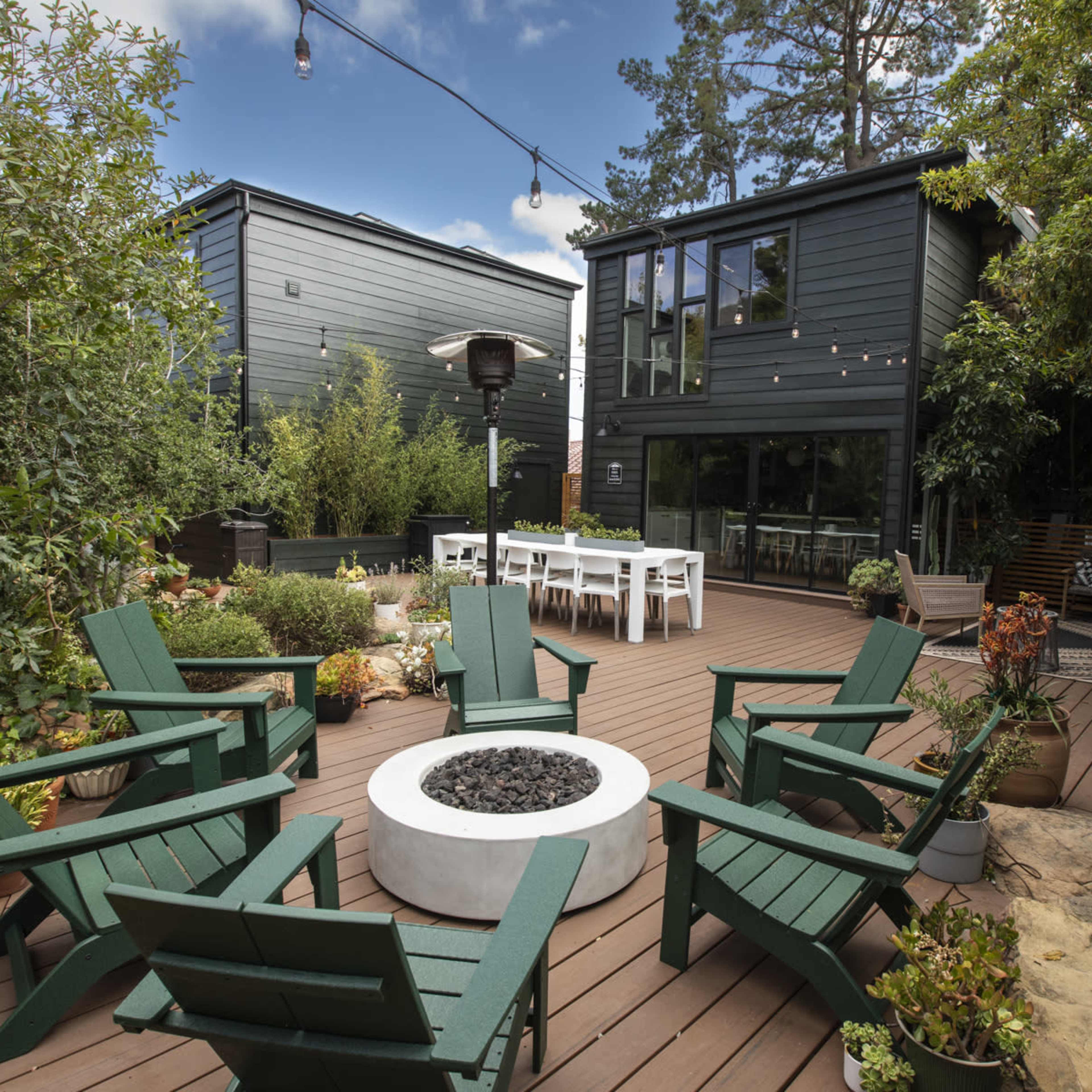 A modern outdoor patio with a fire pit surrounded by green Adirondack chairs, a dining area, and landscaped greenery.