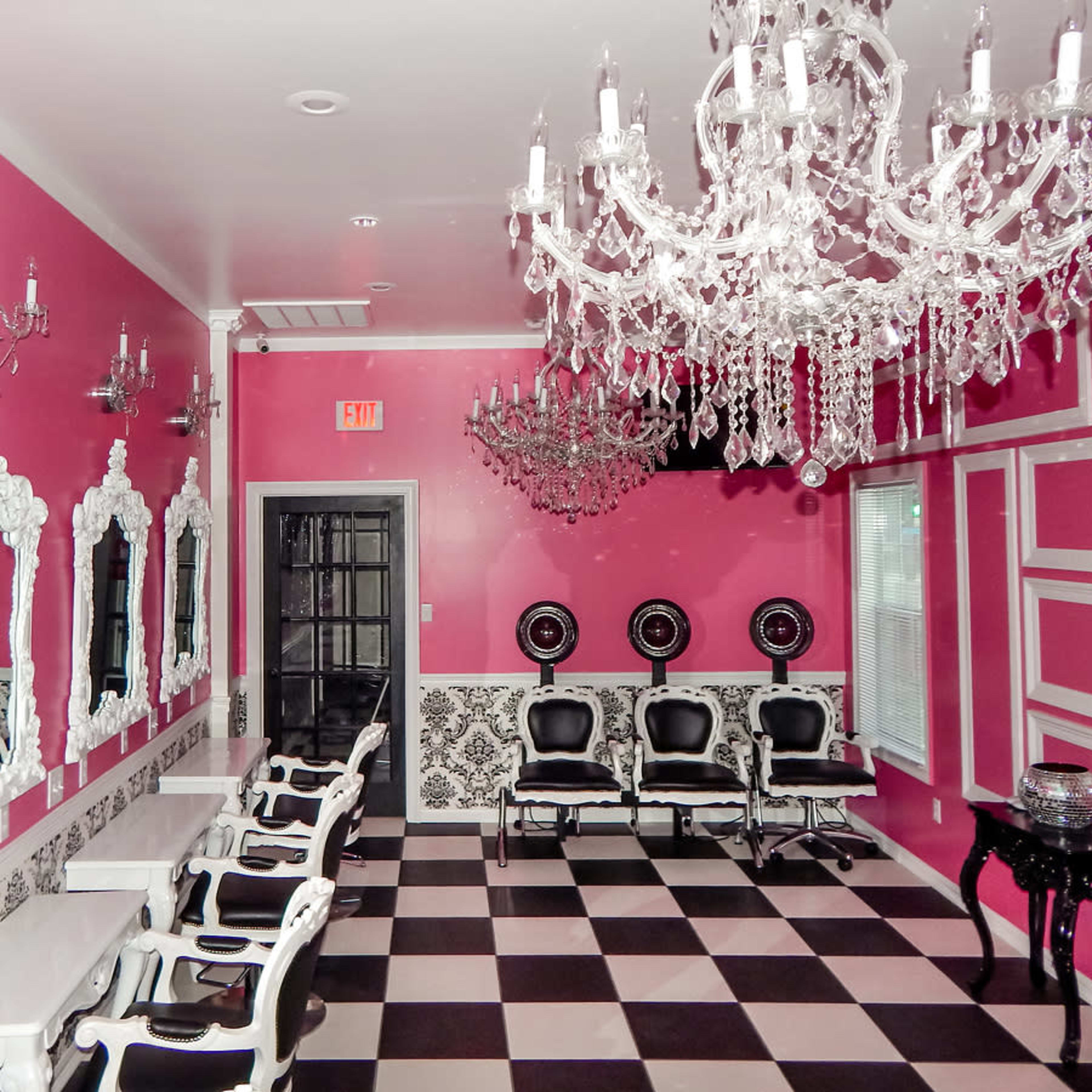 A salon with pink walls, white ornate mirrors, black and white checkered flooring, and chandeliers hanging from the ceiling.