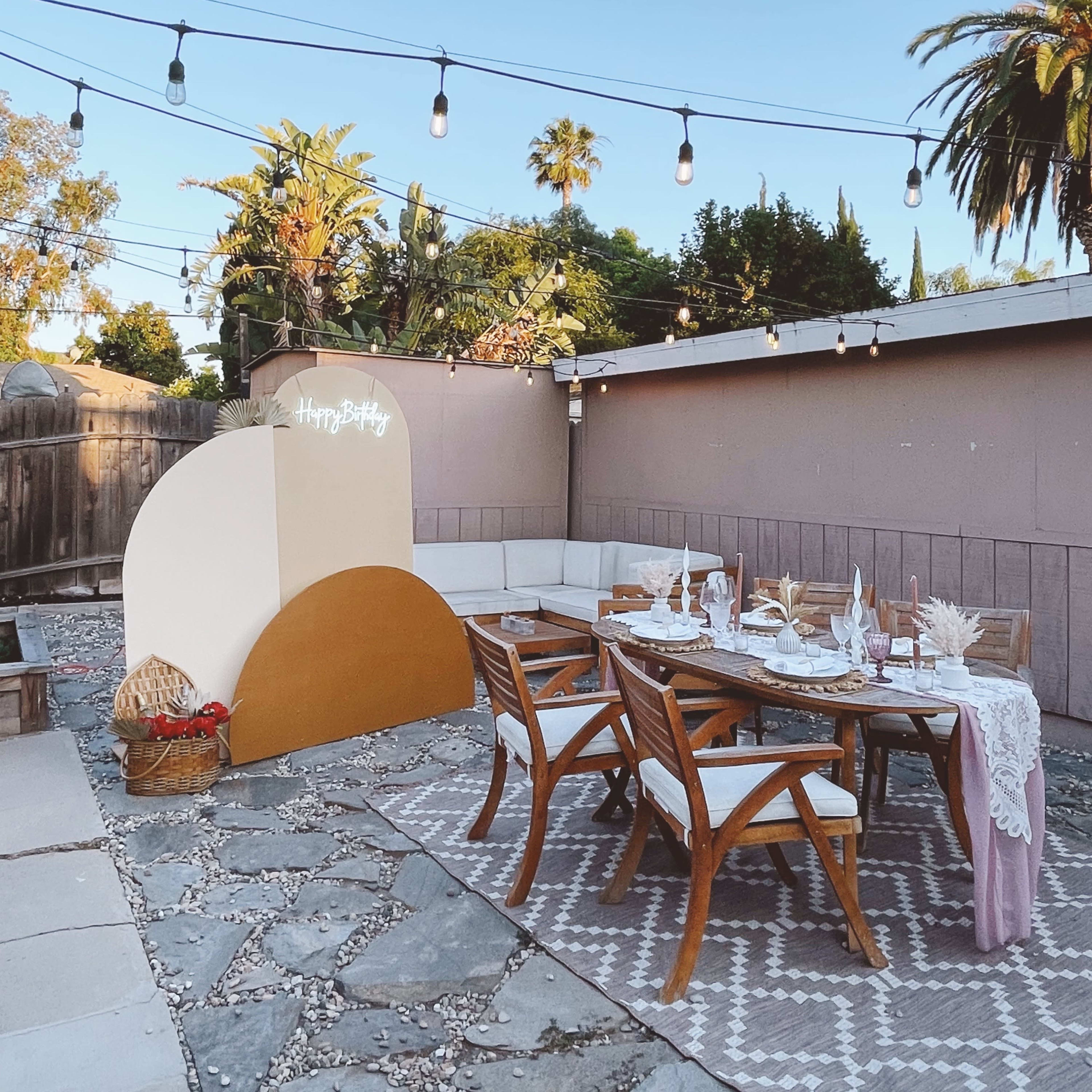 A decorated outdoor dining area features a wooden table set with plates and glasses, surrounded by a stone patio and palm trees, with string lights overhead.