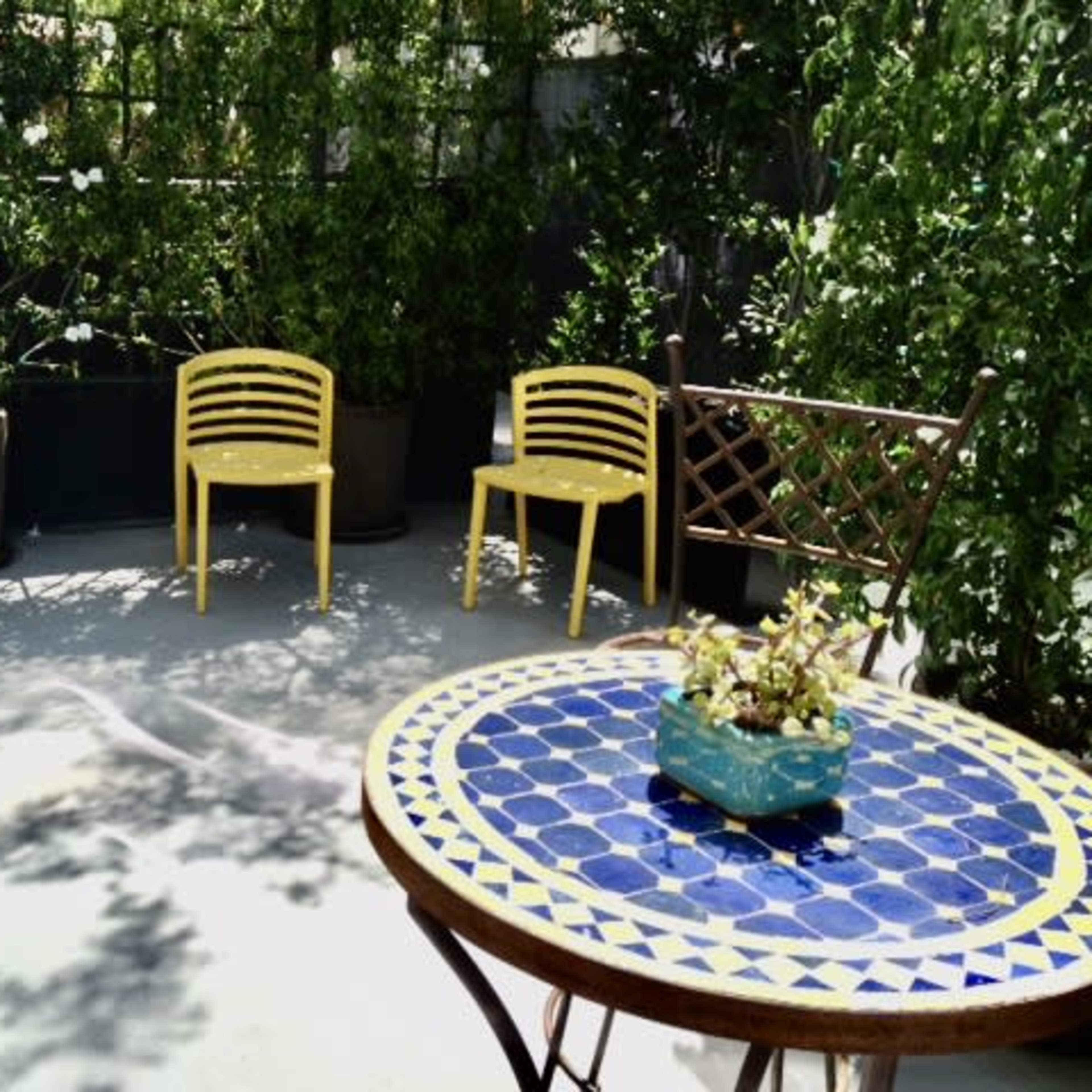 A small outdoor seating area with two yellow chairs, a round table with a blue patterned top, and potted plants surrounded by greenery.