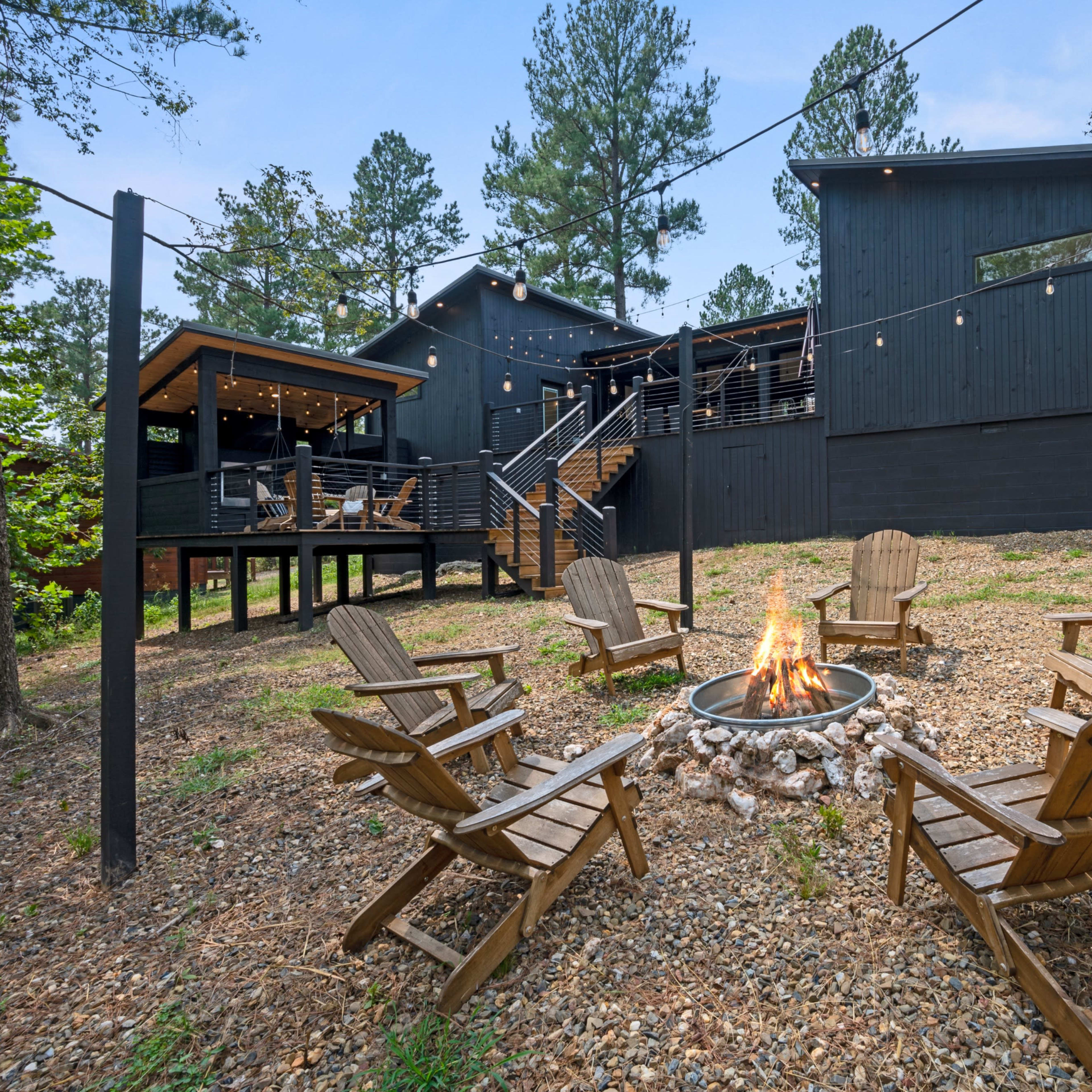 A modern black house is set among trees, featuring a fire pit surrounded by wooden chairs, with string lights overhead and a gravel pathway leading to it.