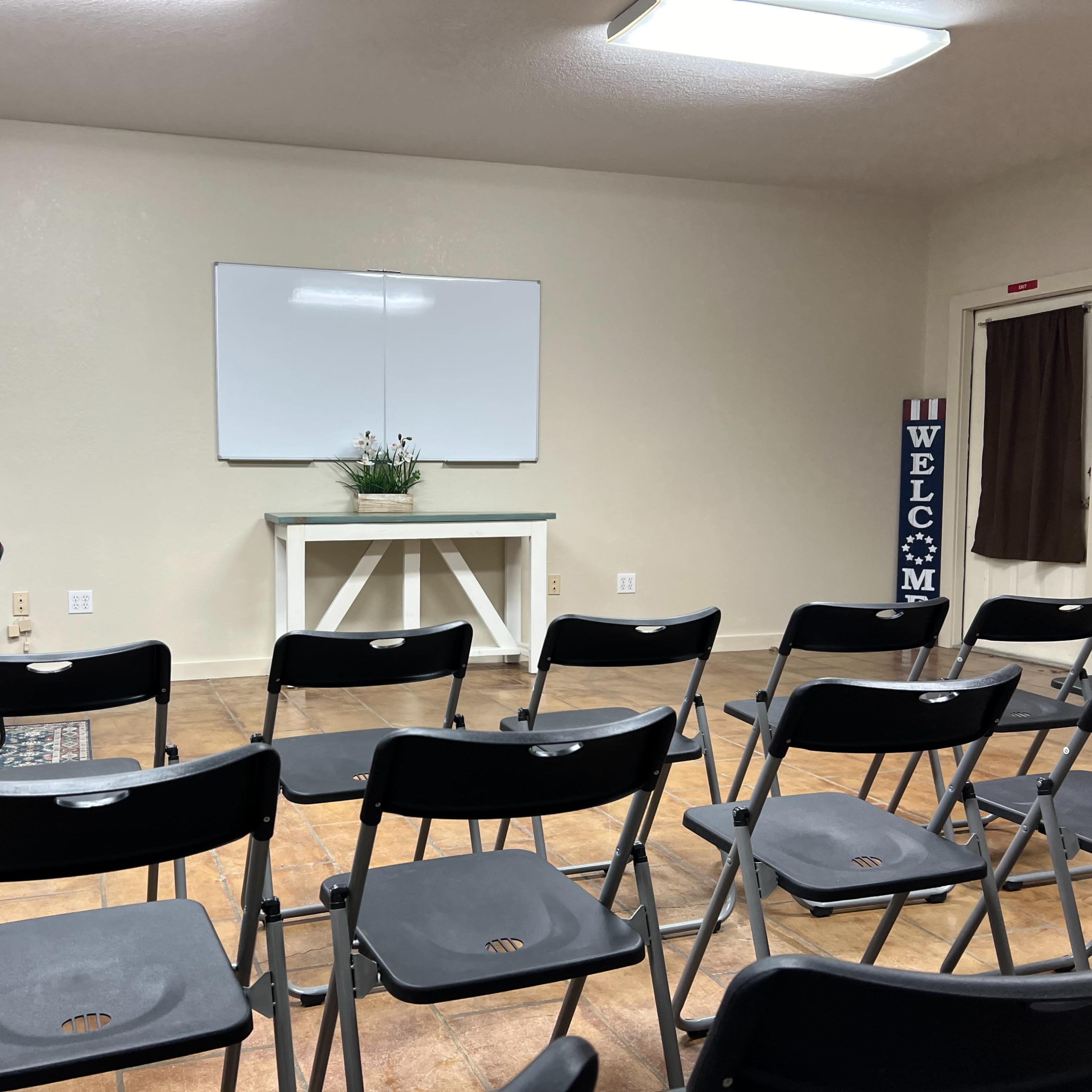 An interior room set up for a meeting, featuring several rows of black folding chairs facing a whiteboard, a table with plants, and a welcome sign.