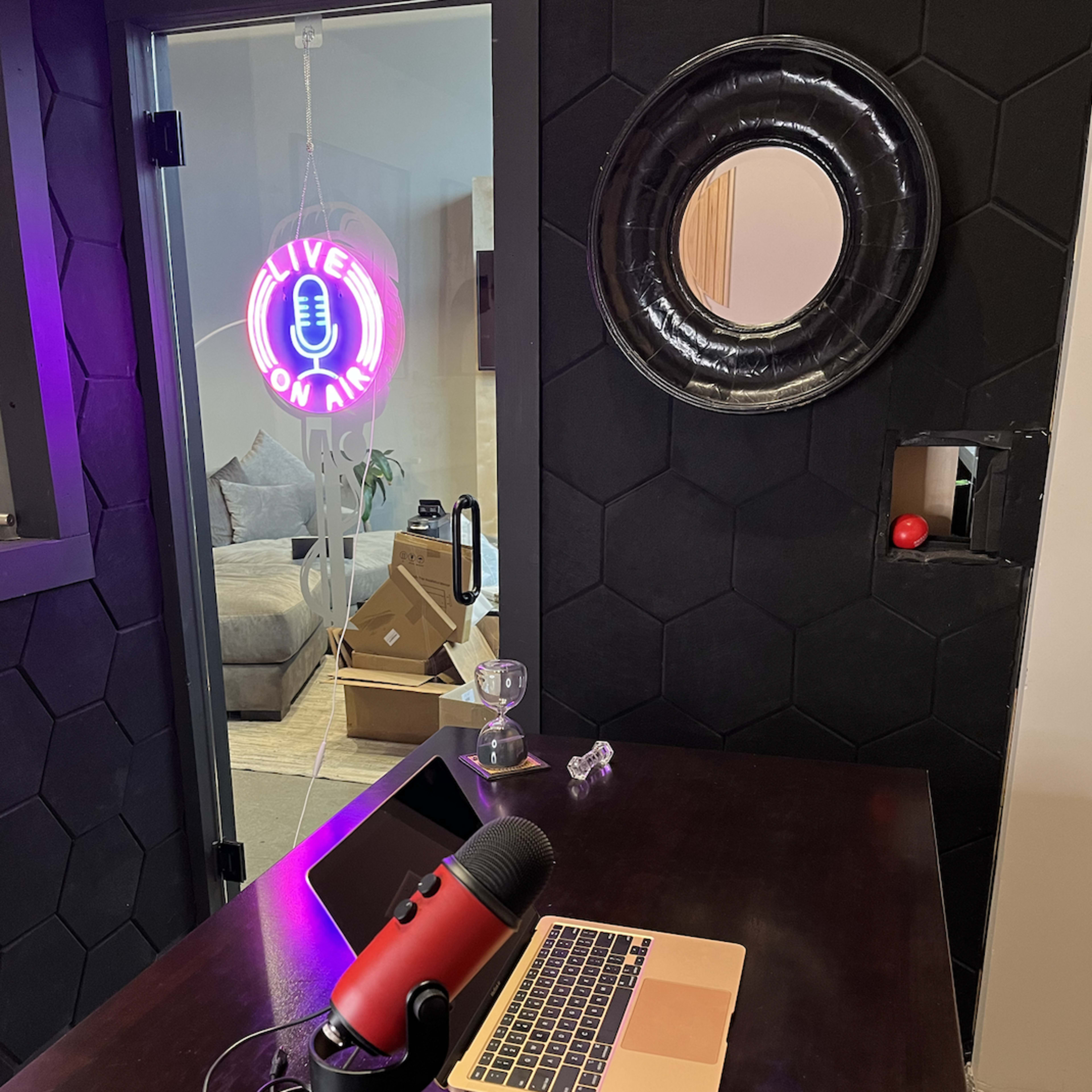 A desk with a red microphone and laptop is positioned in front of a neon "LIVE ON AIR" sign, with a doorway leading to a room in the background.