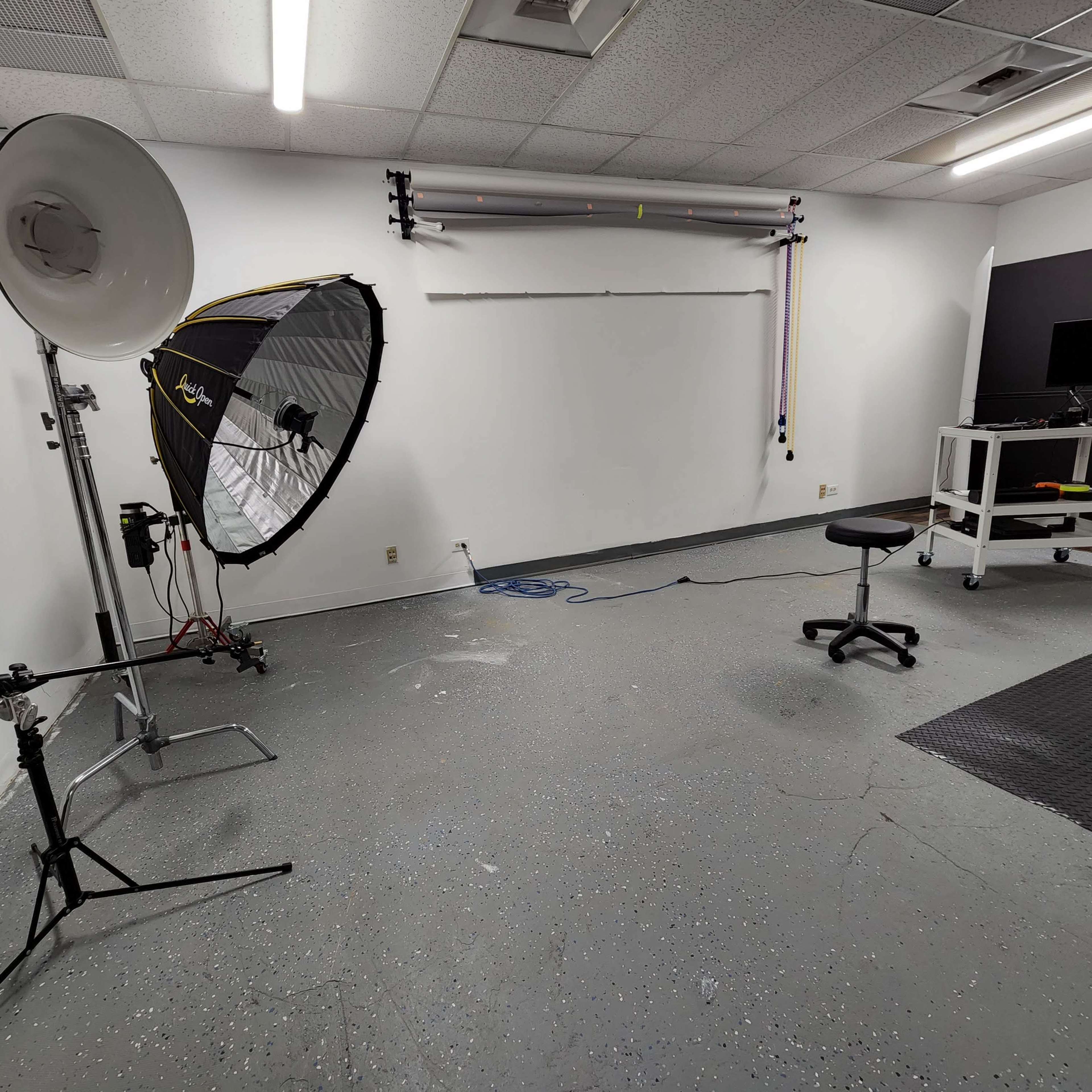 An empty photography studio featuring a large softbox light, a stool, a backdrop support system, and a mobile workstation.