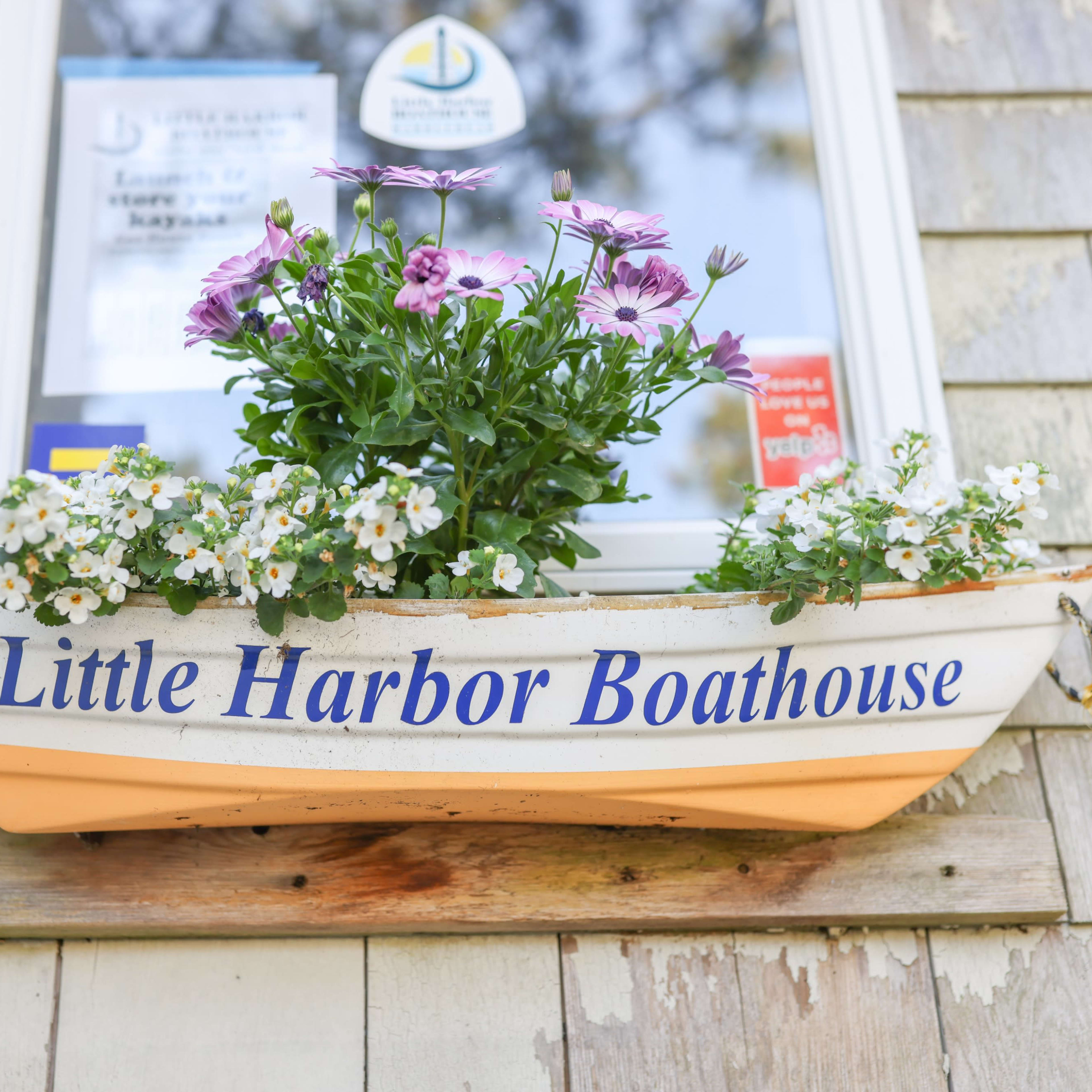 A small, decorative boat filled with colorful flowers is displayed outside a window, labeled "Little Harbor Boathouse."