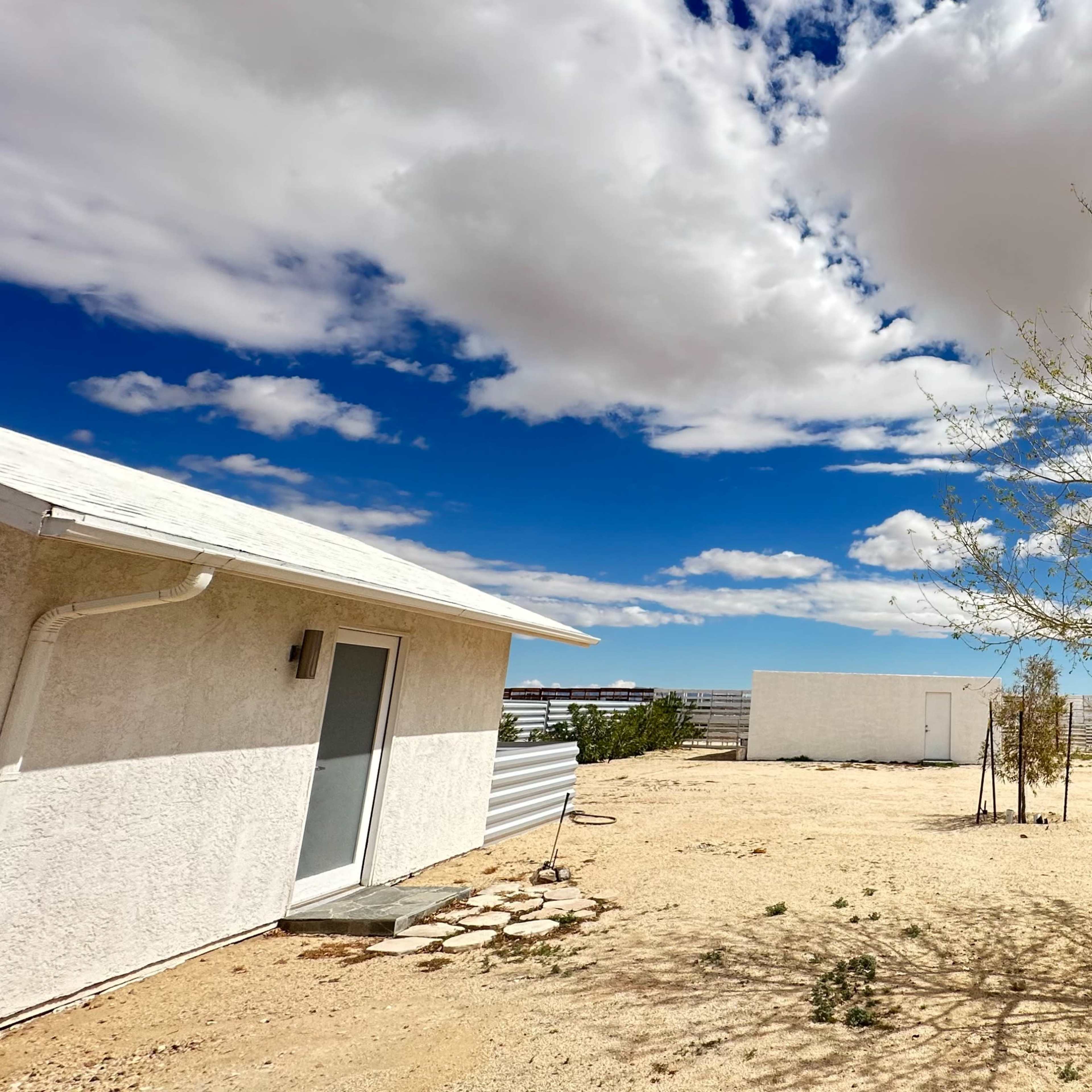A white building and a shed are surrounded by sparse vegetation and a sandy landscape under a blue sky with scattered clouds.