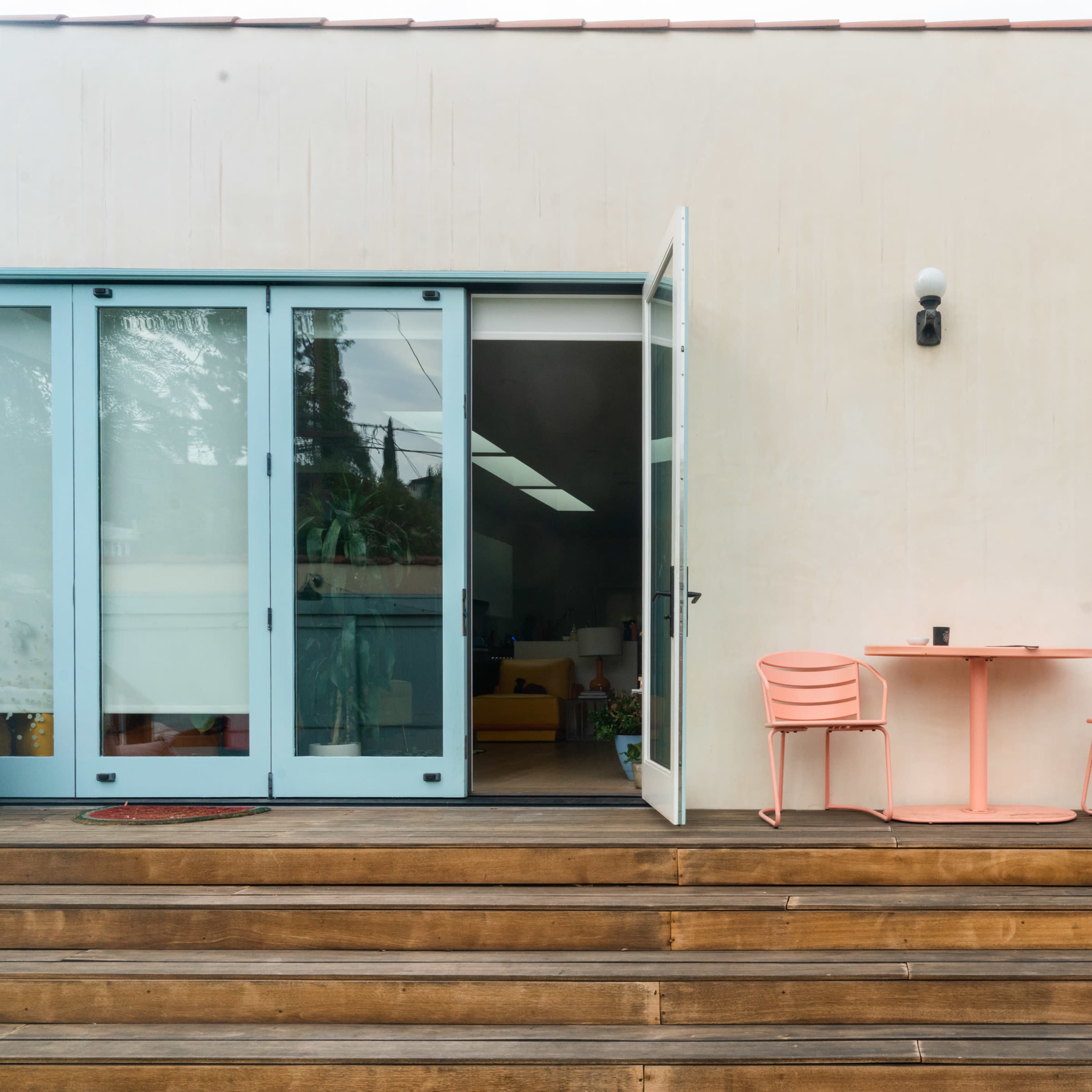 A modern patio with a light-colored building, a small round table, and two pink chairs, all situated on wooden steps.