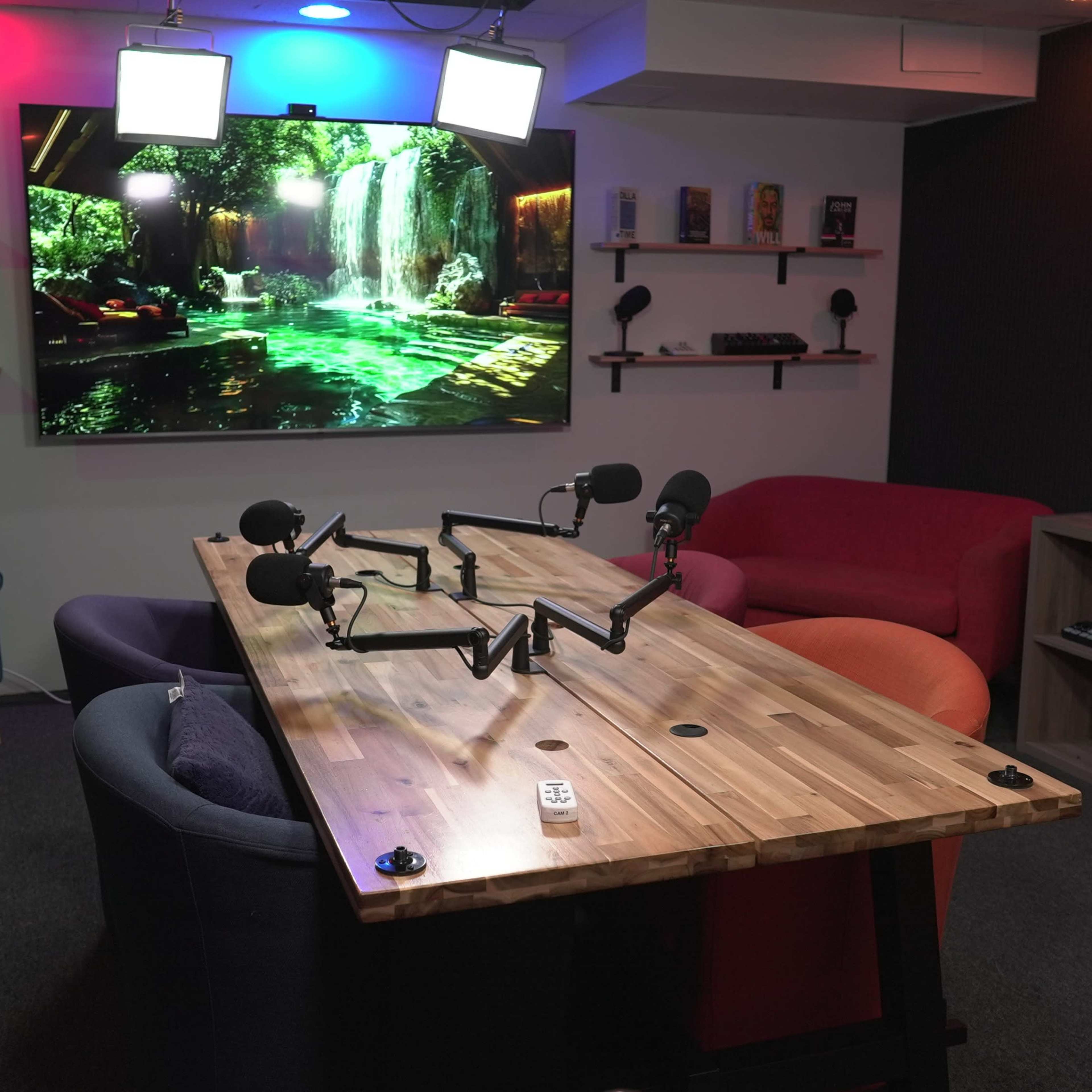 A modern room set up for recording, featuring a large screen displaying a waterfall scene, a wooden table with microphone stands, and colorful seating arrangements.