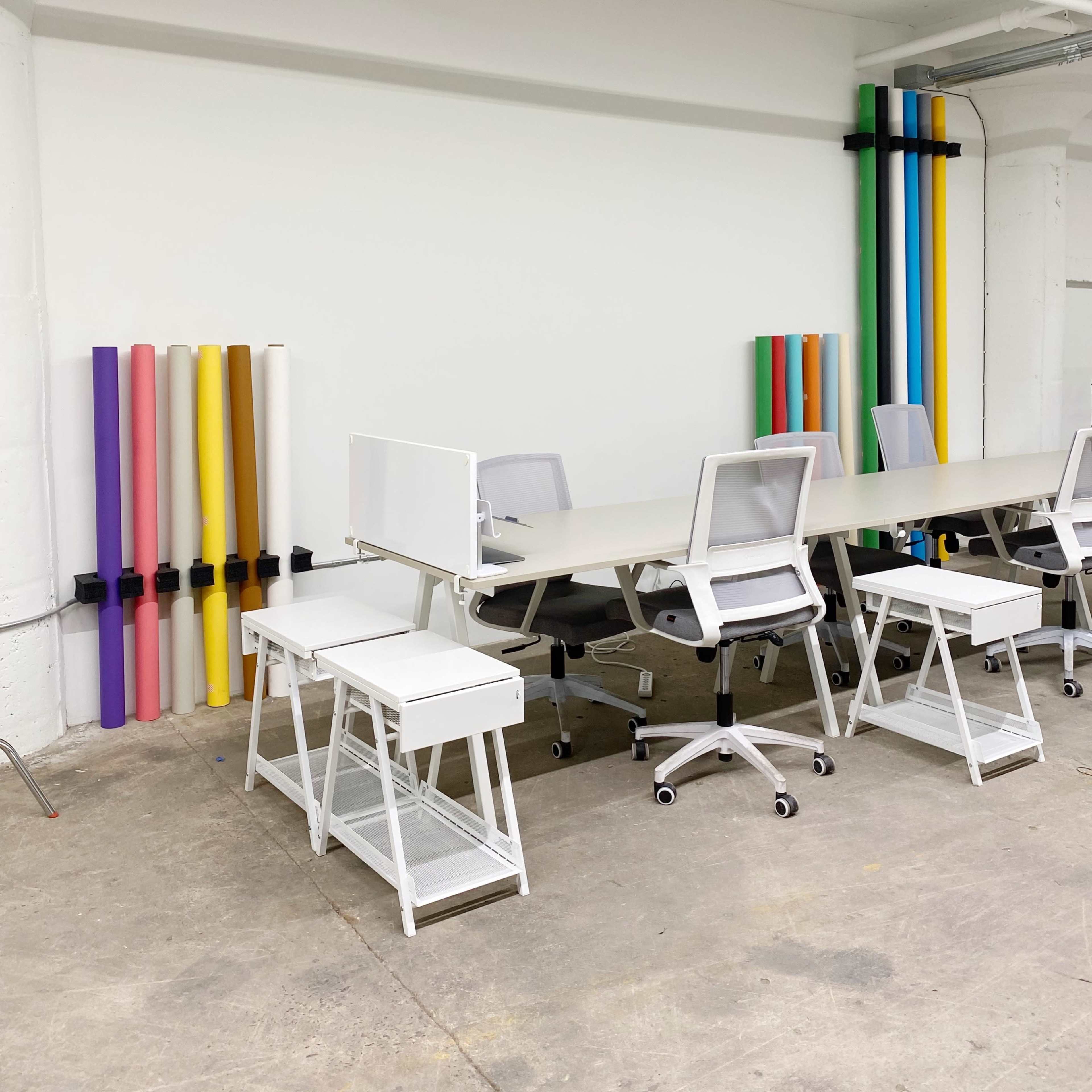 A workspace with several white desks and chairs, complemented by colorful rolls of paper stacked vertically against the wall.