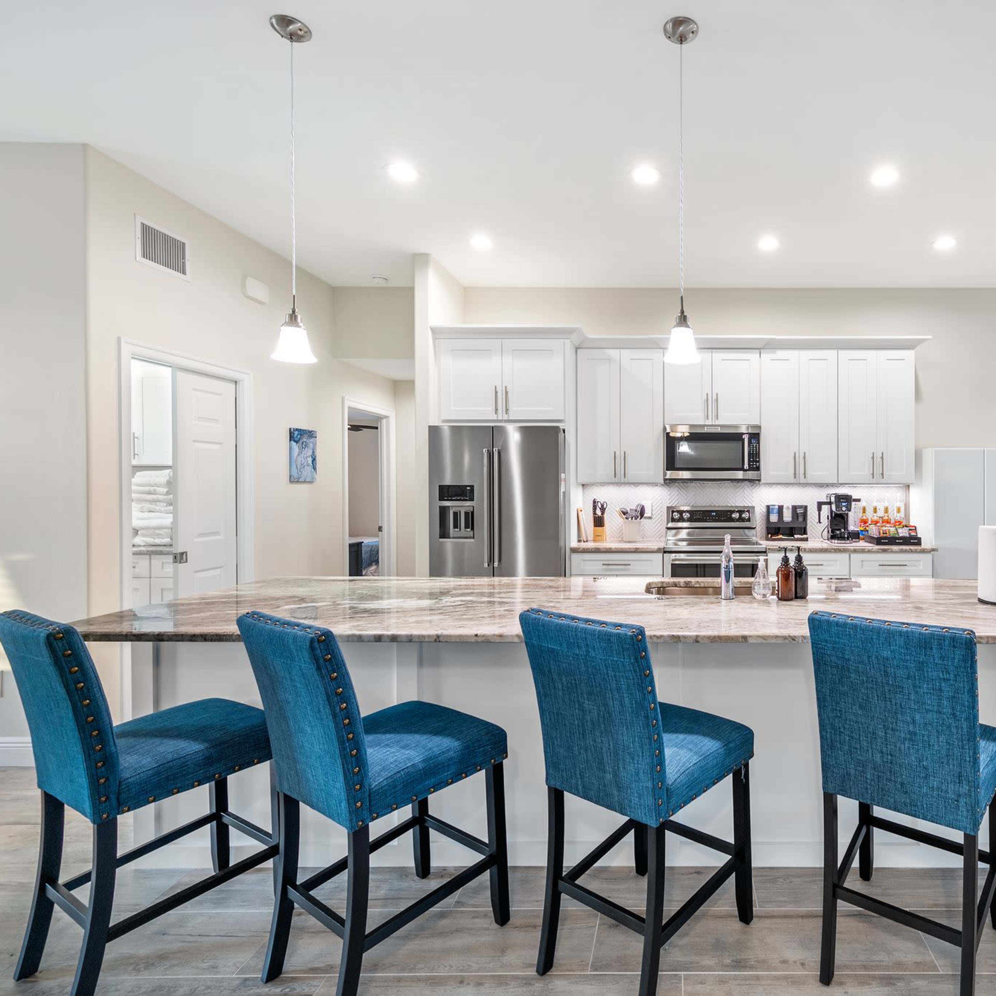 A modern kitchen features a large island with four blue barstools, stainless steel appliances, and white cabinetry.