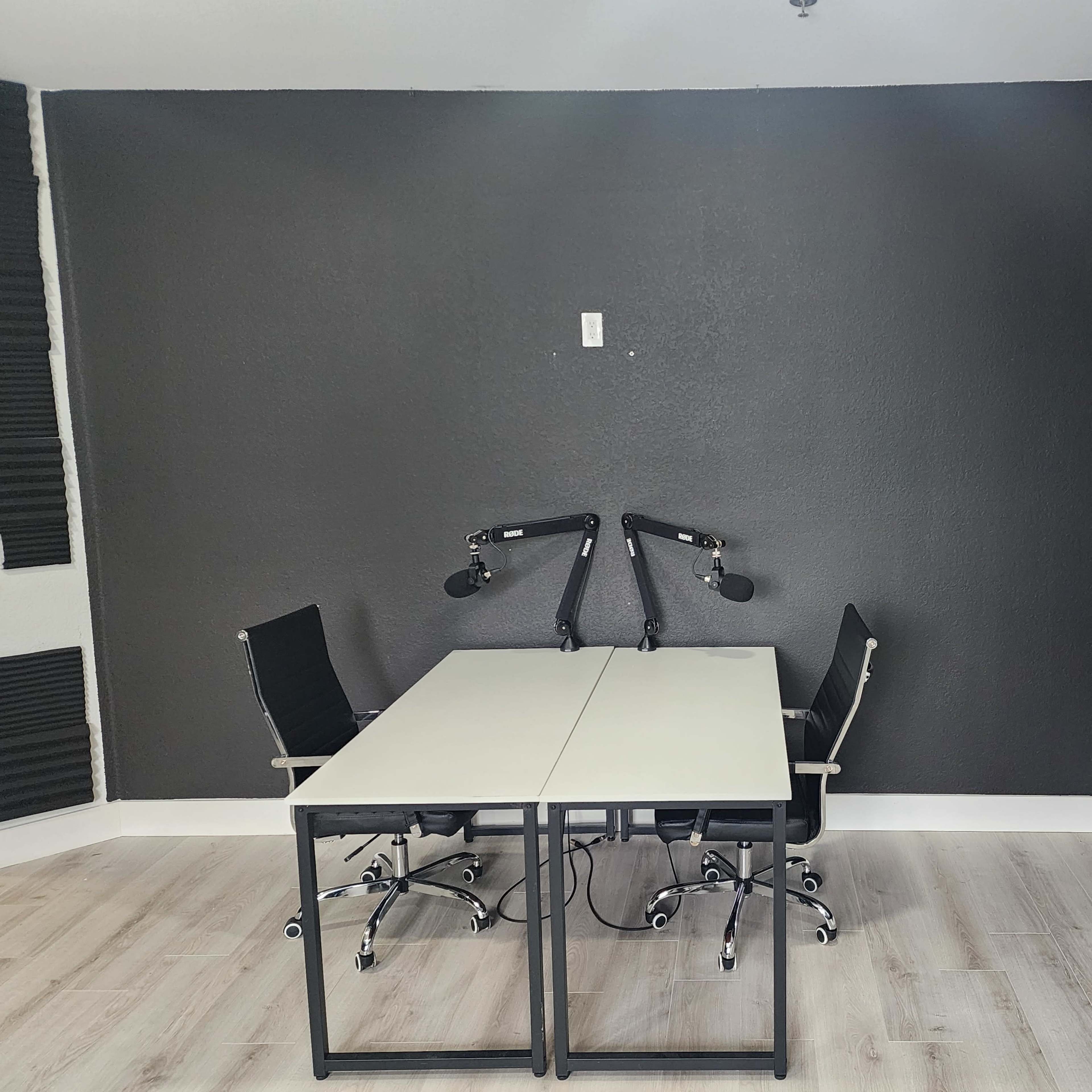 A room with a black accent wall, two chairs with armrests at a long table, and mounted microphone stands on the table.