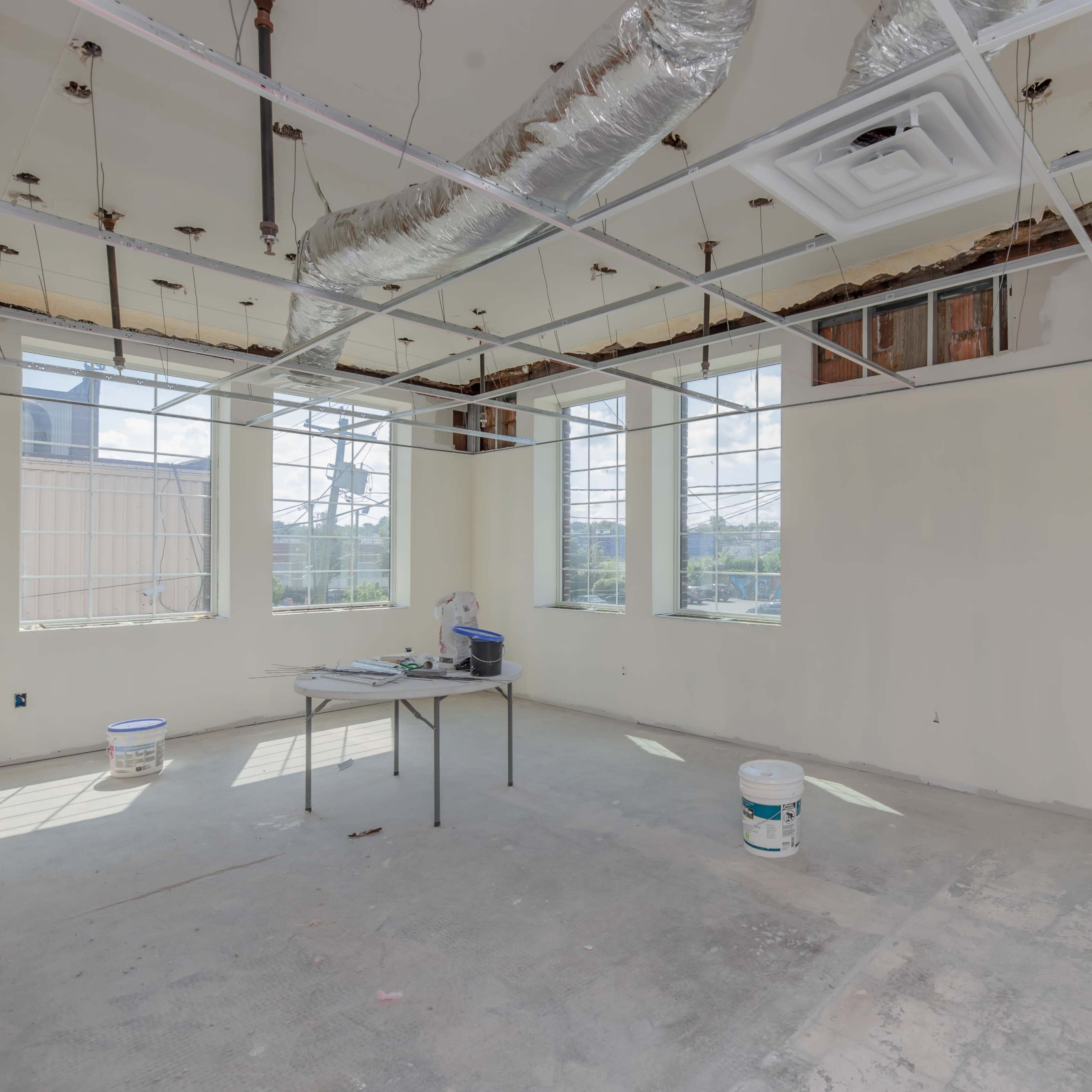An empty room with unfinished walls, exposed ceiling structures, and large windows allowing natural light to enter.