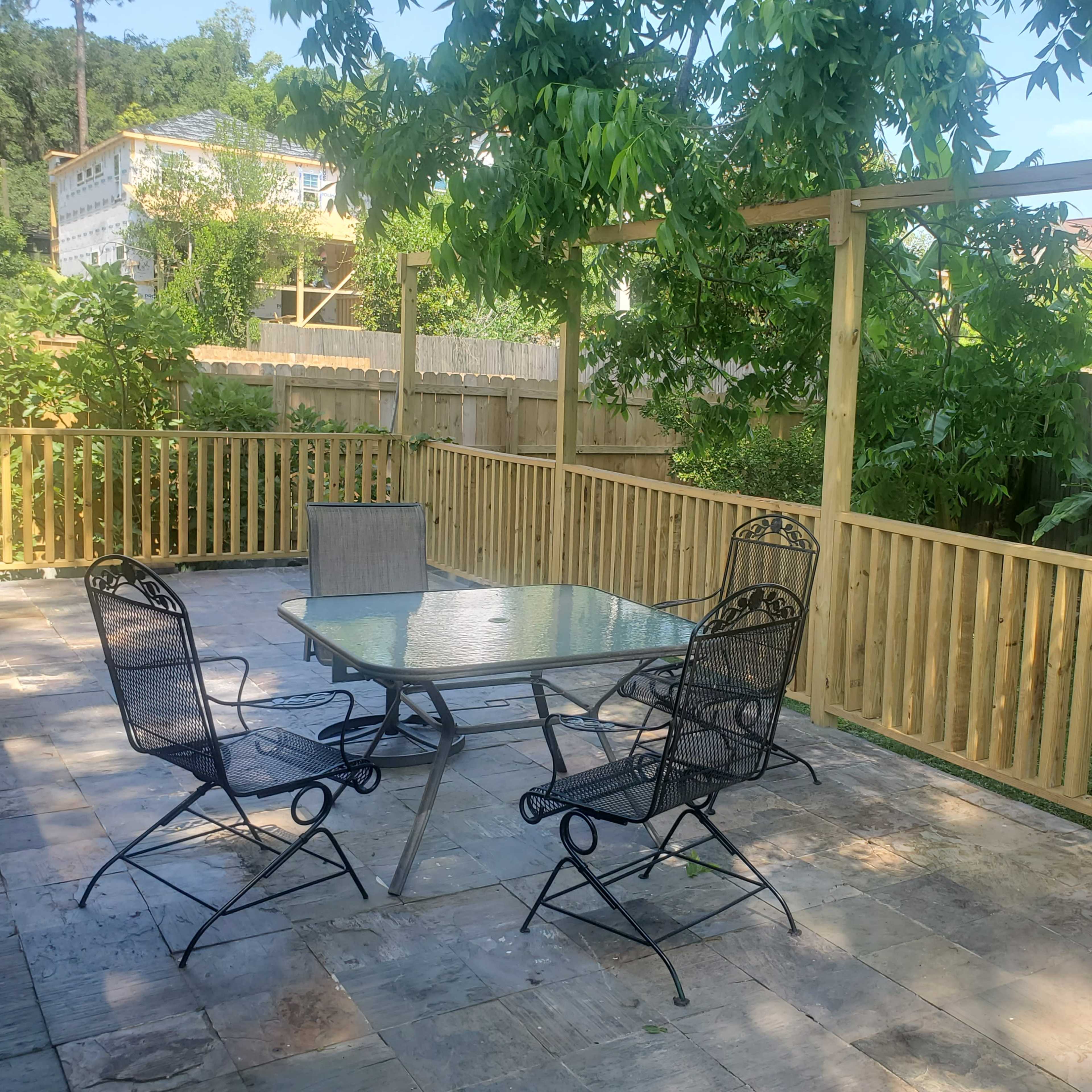 A patio with a glass table surrounded by four black metal chairs is enclosed by a newly built wooden fence and greenery.
