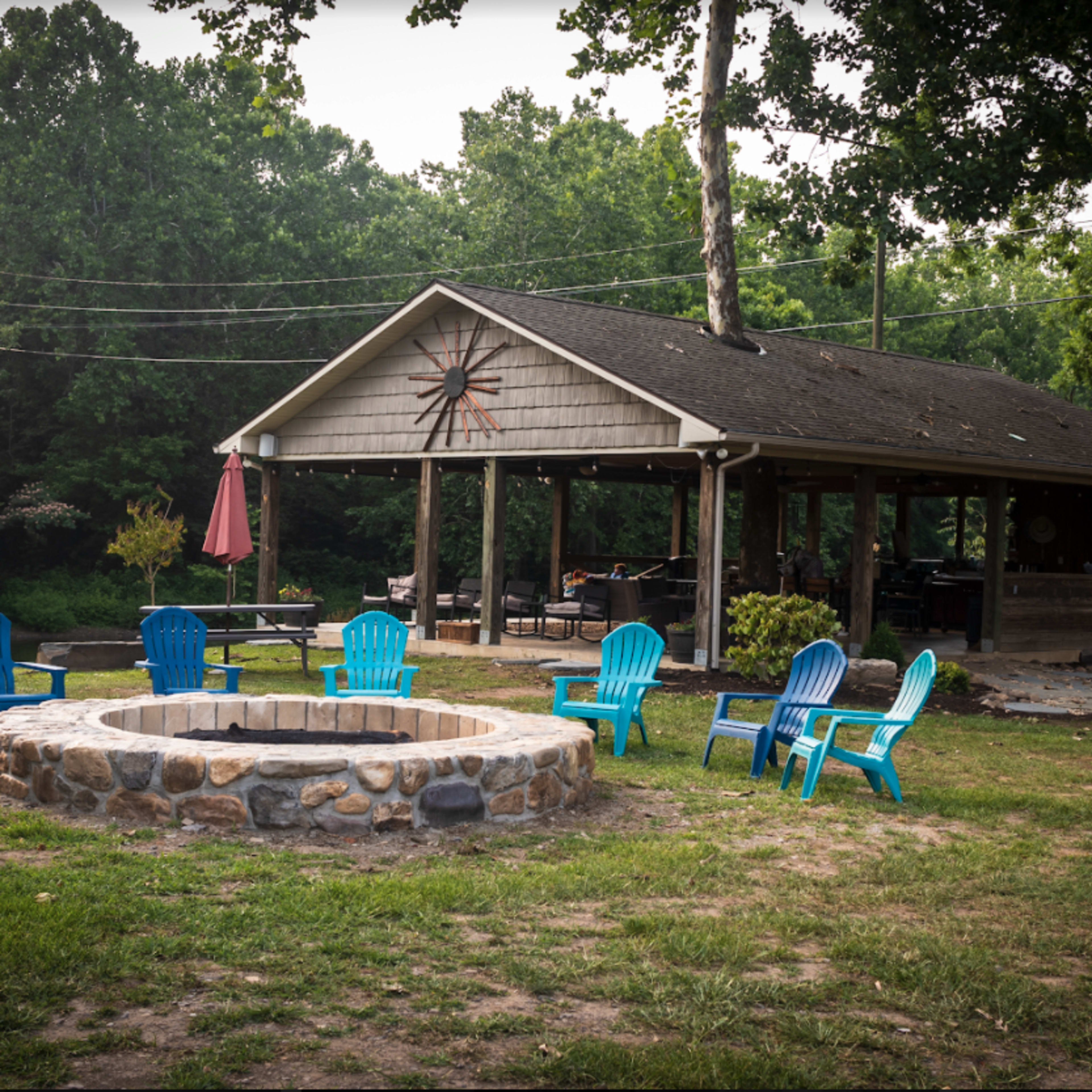 A stone fire pit surrounded by blue adirondack chairs is located in front of a wooden pavilion set in a wooded area.