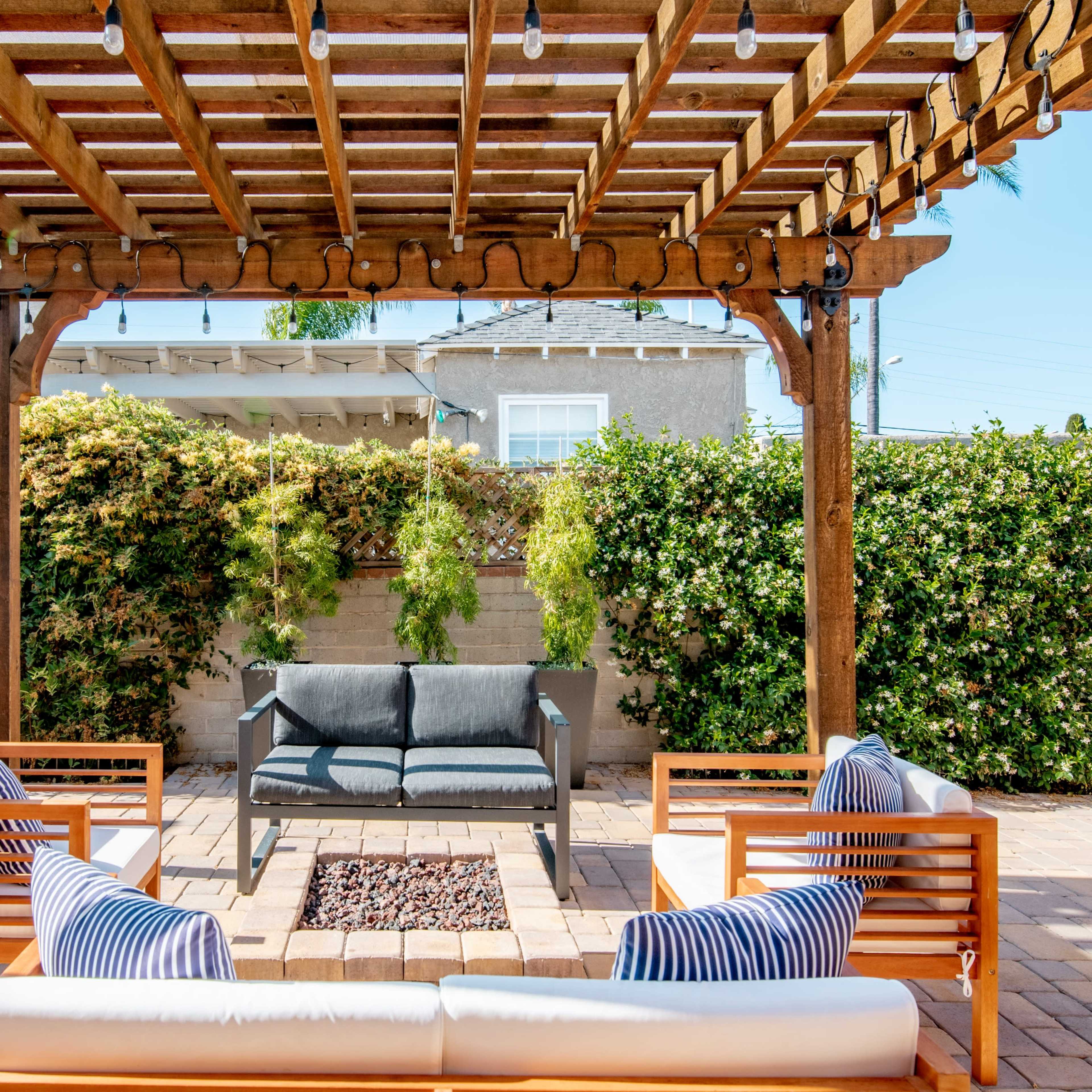 A covered patio with wooden beams features a seating area arranged around a fire pit, surrounded by lush greenery.