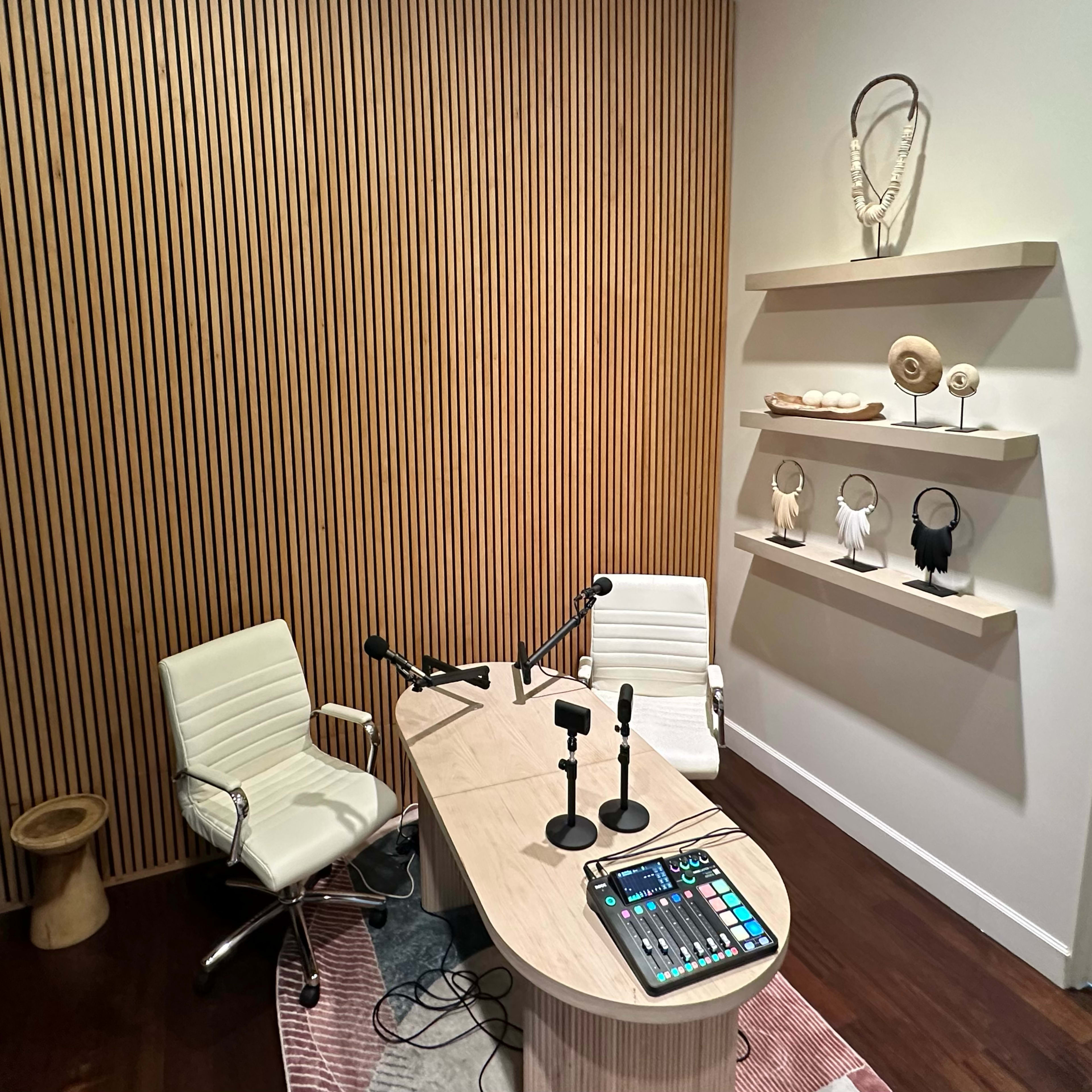 A modern recording studio featuring a wooden desk with two microphones, a sound mixing panel, and decorative shelves displaying various objects against a wall with vertical wooden paneling.