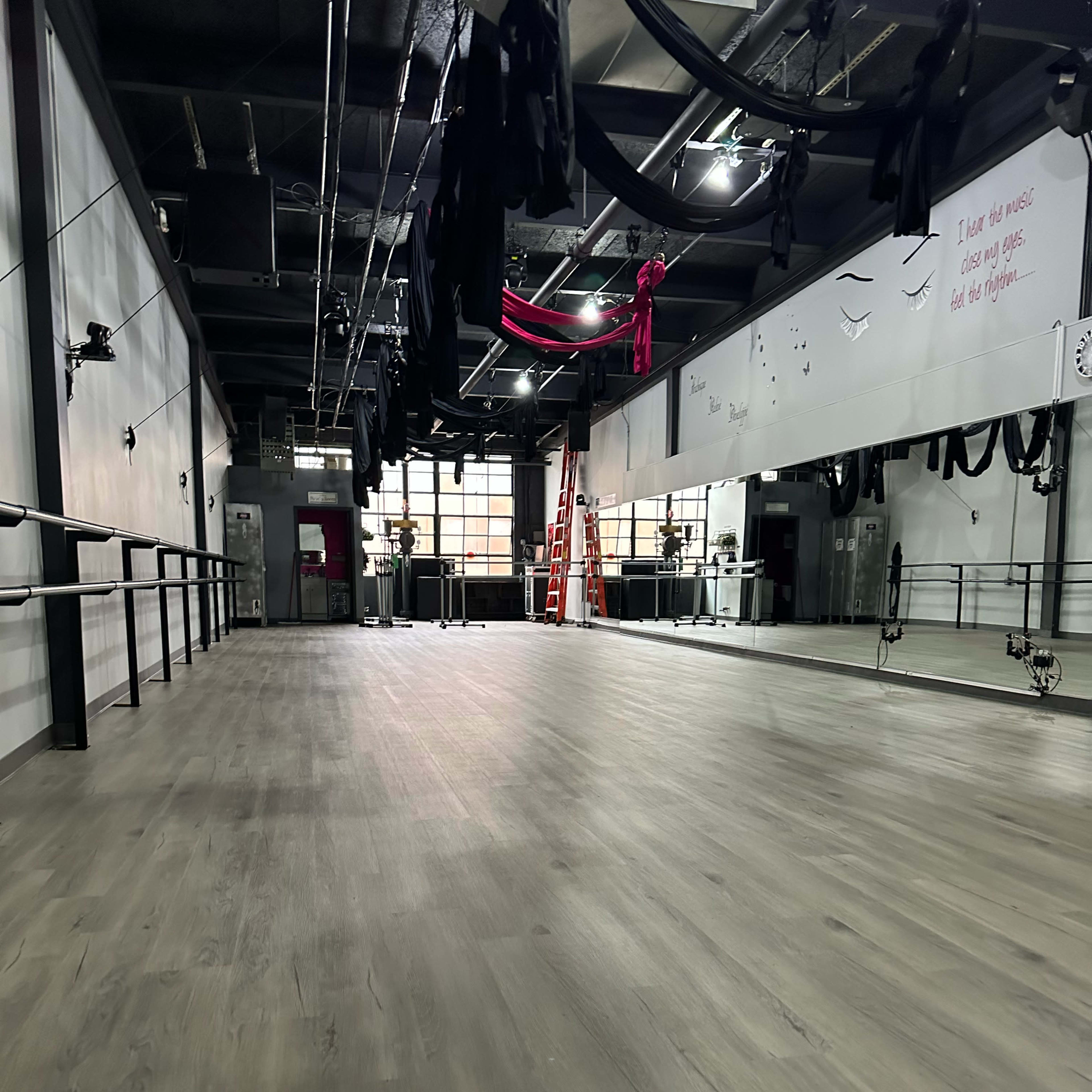 A spacious dance studio with wooden floors, mirrors along the wall, and dance equipment suspended from the ceiling.