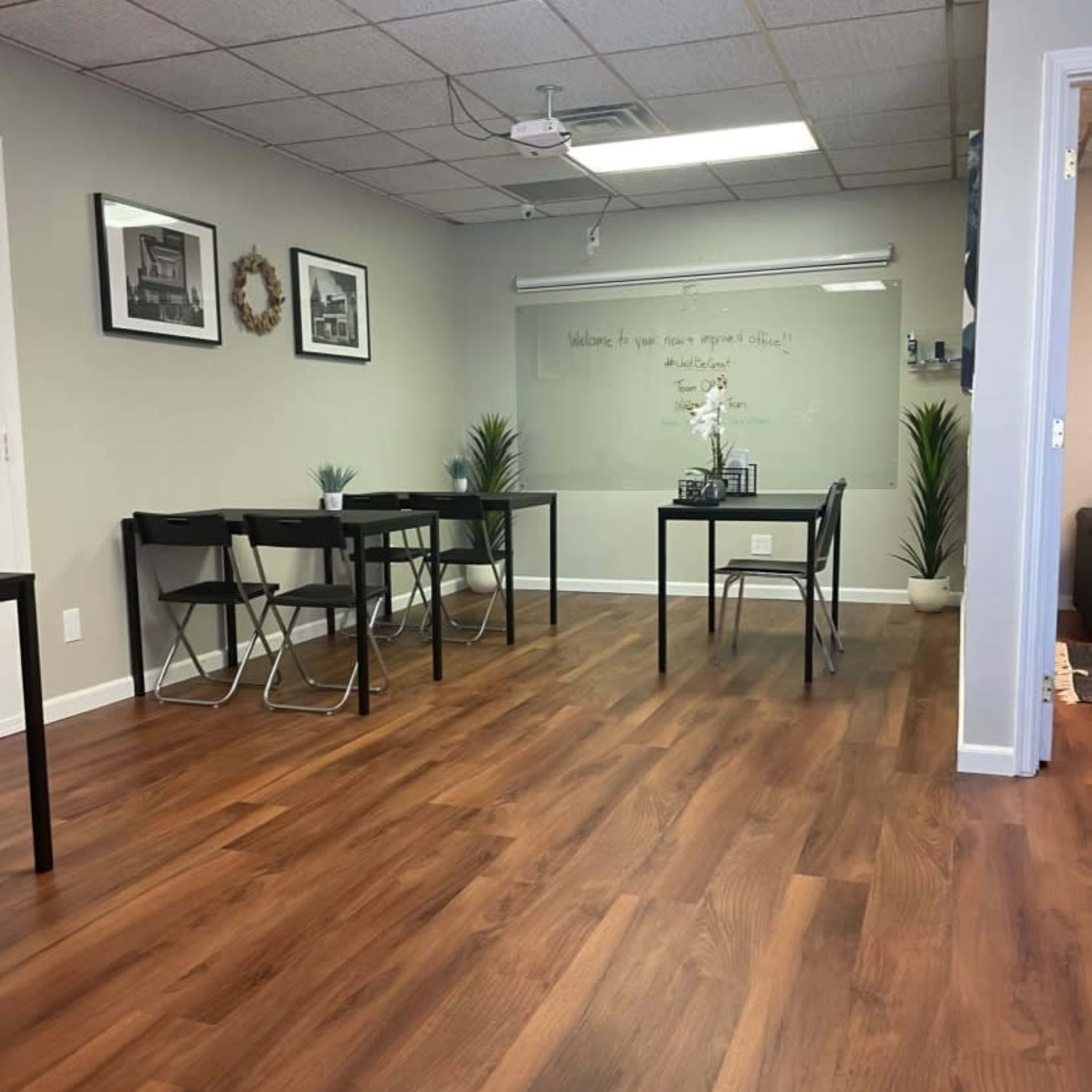 A spacious room with wooden flooring, featuring several black tables and chairs, along with wall decorations and plants.