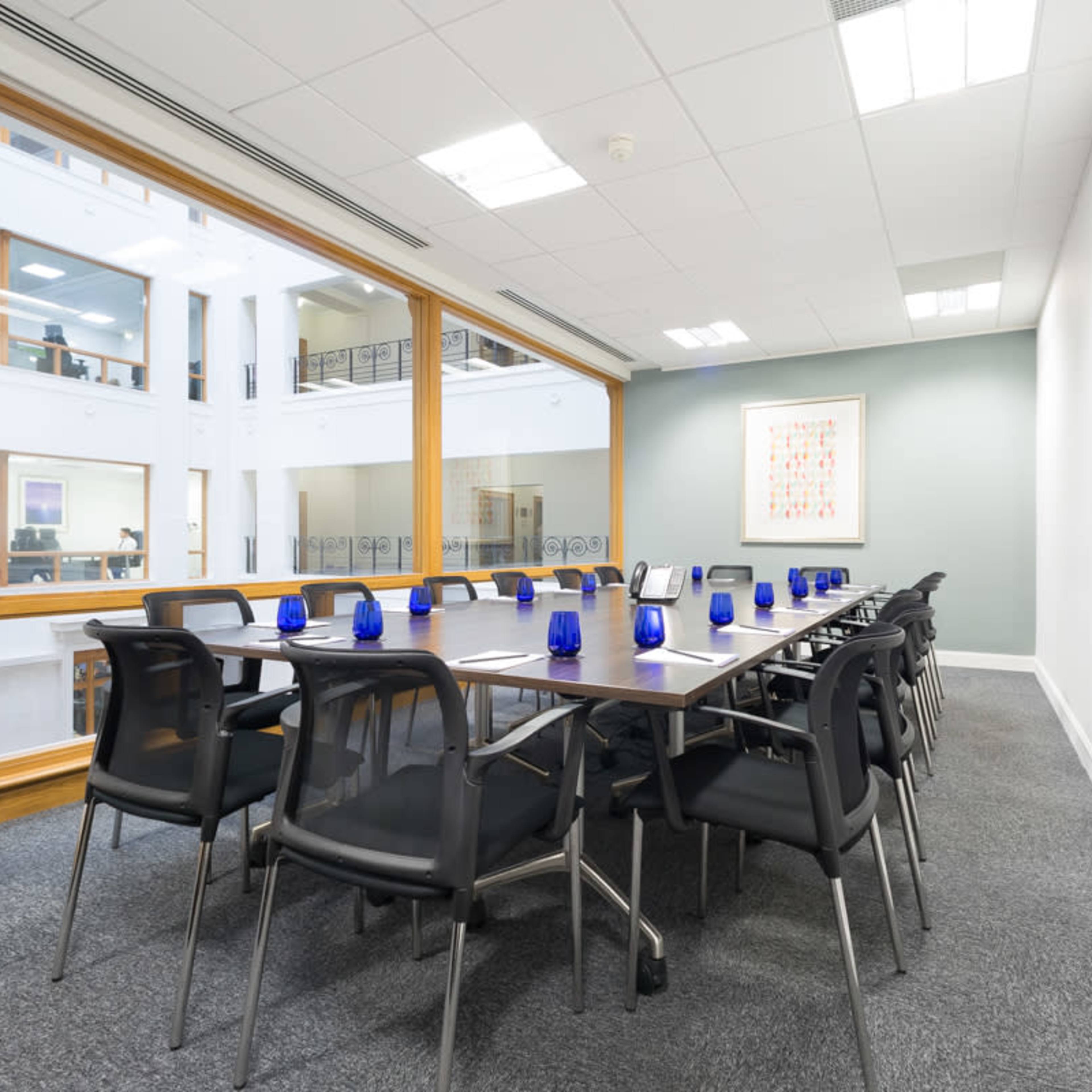 A long conference table with black chairs is arranged in a bright meeting room featuring large windows and blue glass decor.